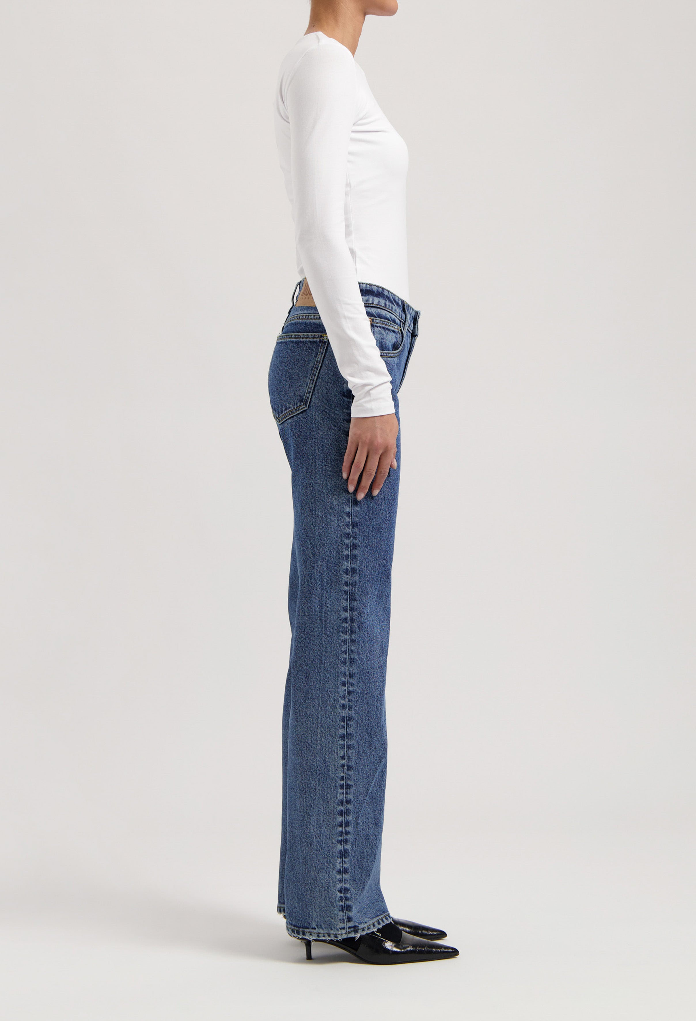 Side profile of a person wearing classic blue straight-leg jeans paired with a fitted white long-sleeve top and black heels.