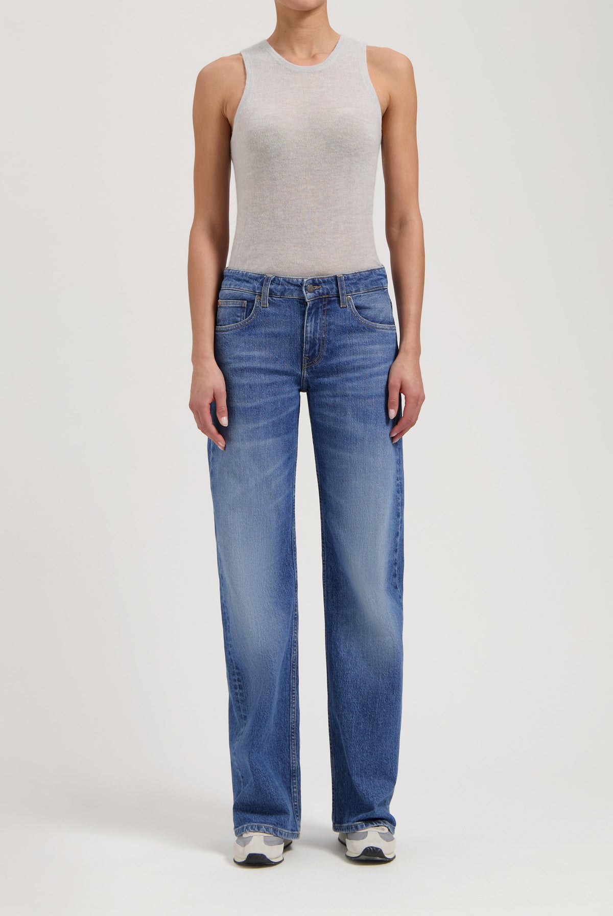 Woman wearing gray sleeveless top and relaxed-fit blue denim jeans, paired with white sneakers against a plain background.