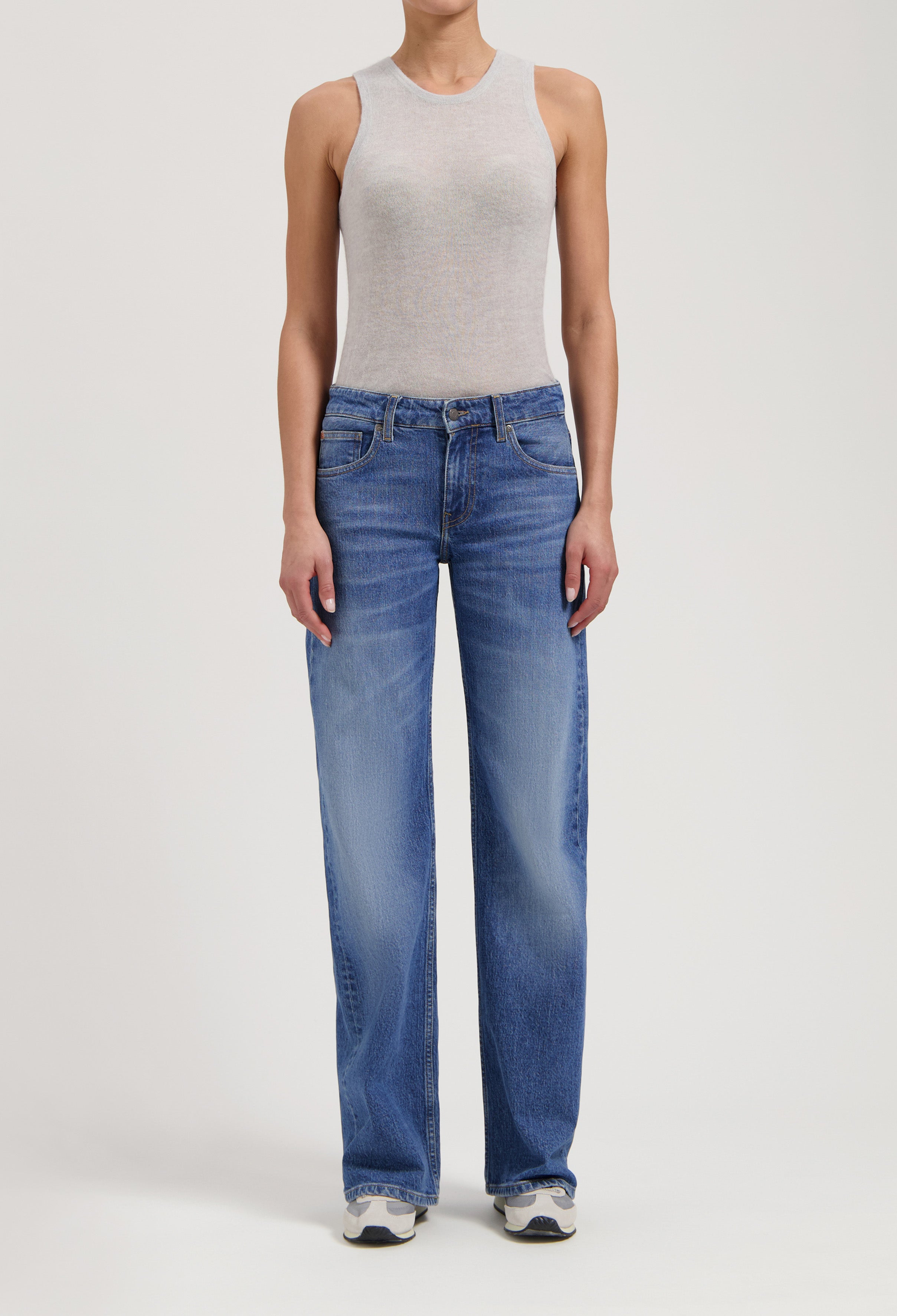 Woman wearing gray sleeveless top and relaxed-fit blue denim jeans, paired with white sneakers against a plain background.