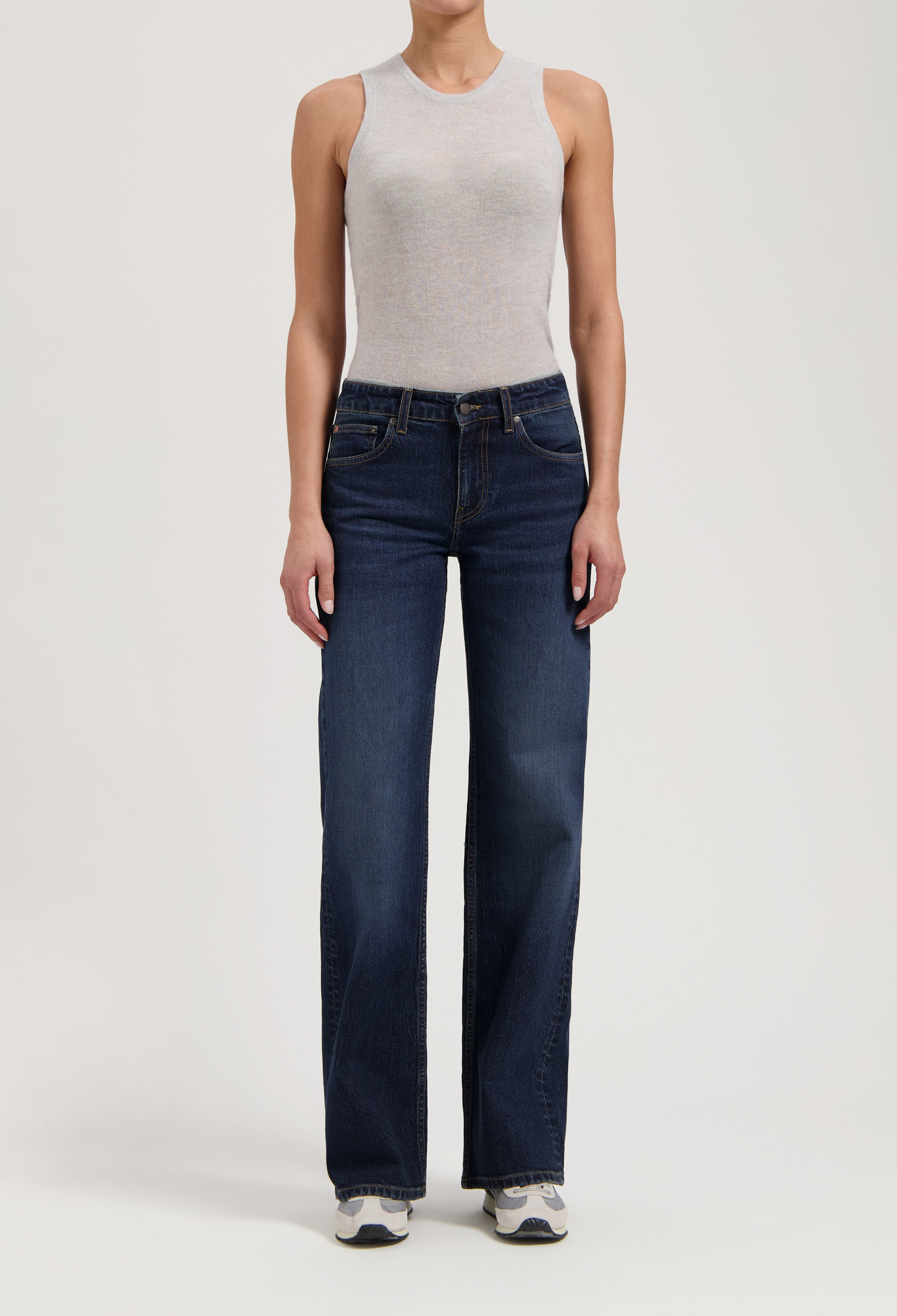 Woman wearing a light gray sleeveless top paired with dark blue, wide-leg jeans and white sneakers, standing in studio.