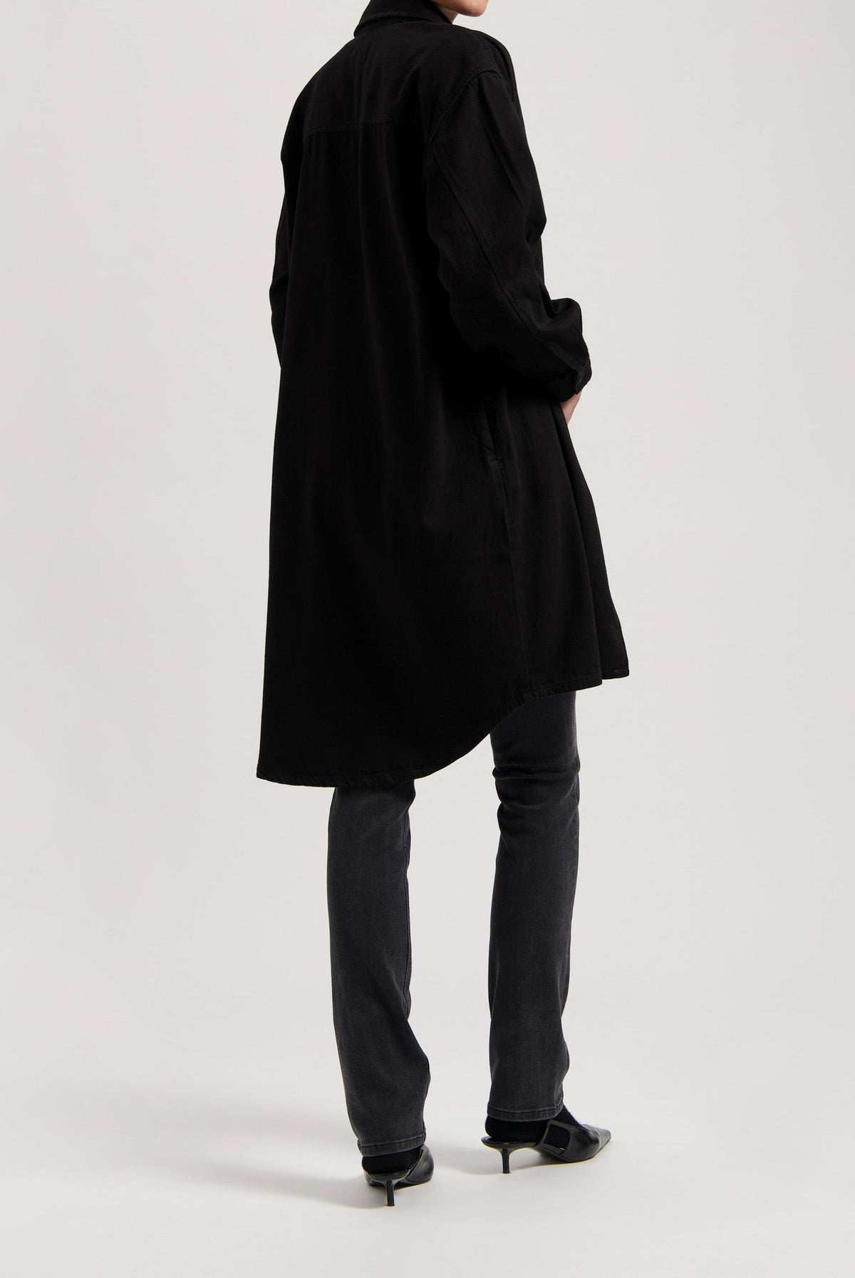 Long black shirt dress styled over dark denim jeans and black pointed-toe heels, showcasing a minimalist fashion look.