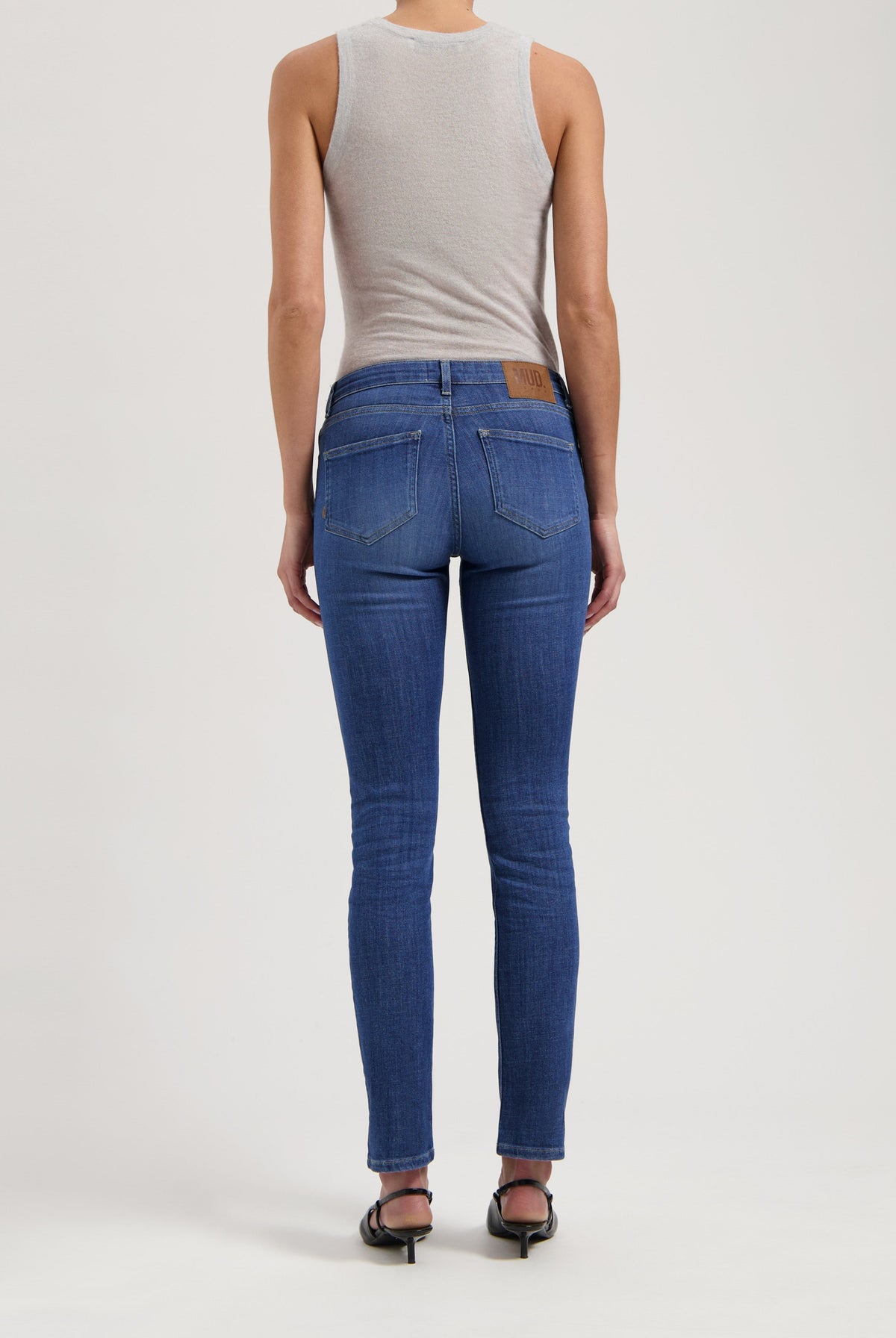 Back view of slim-fit, sustainable blue jeans by MUD Jeans, paired with a light gray sleeveless top and black sandals.