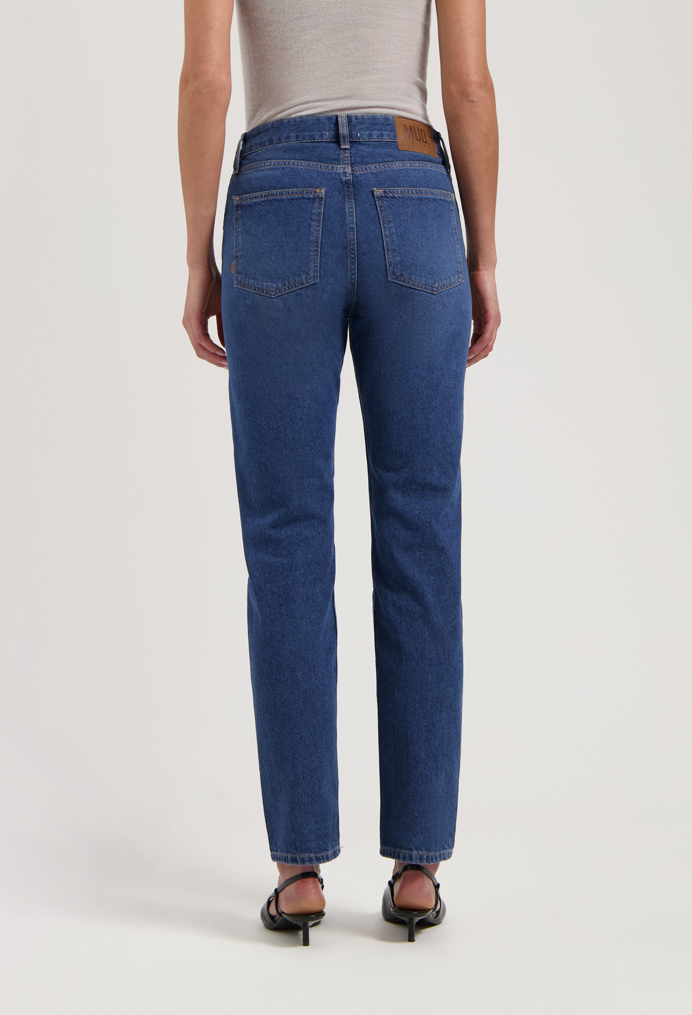 Rear view of sustainable MUD Jeans in dark blue, straight-fit denim worn with a beige tank top and black sandals.