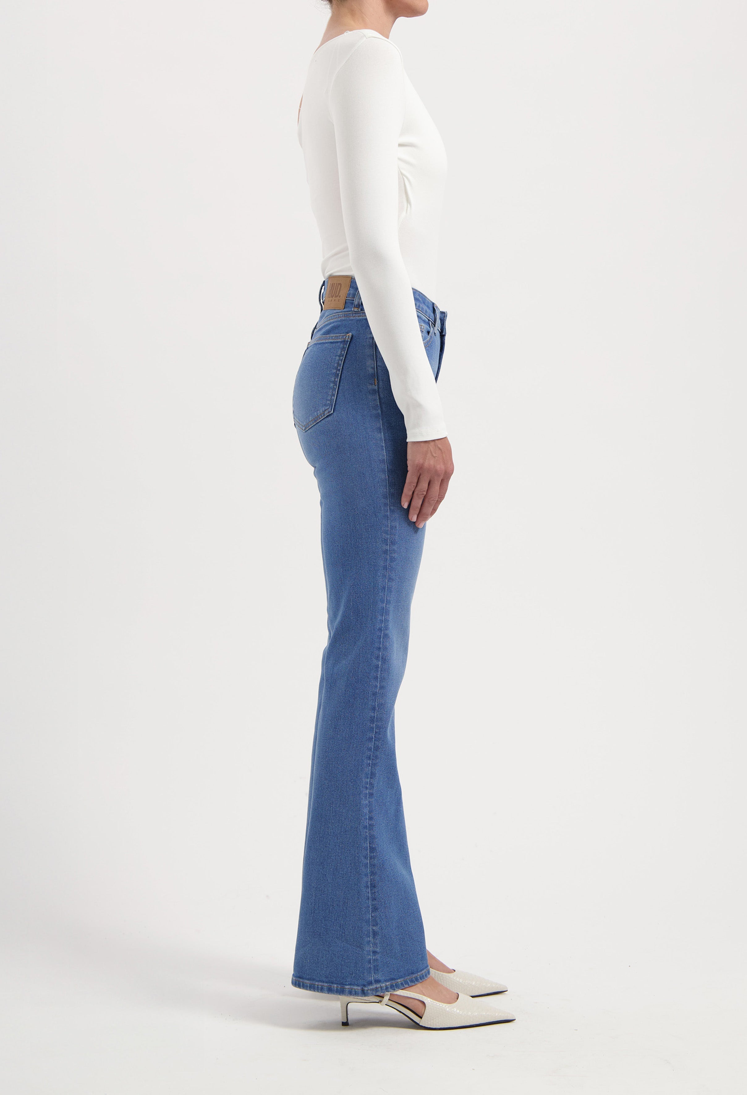 Model wearing light blue, high-waisted flared jeans paired with a fitted white long-sleeve top and cream pointed heels.