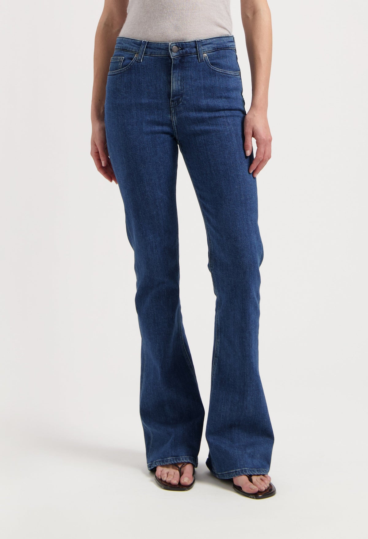 Woman wearing bootcut blue denim jeans, showcasing a classic fit, paired with a beige sleeveless top and flip-flops.