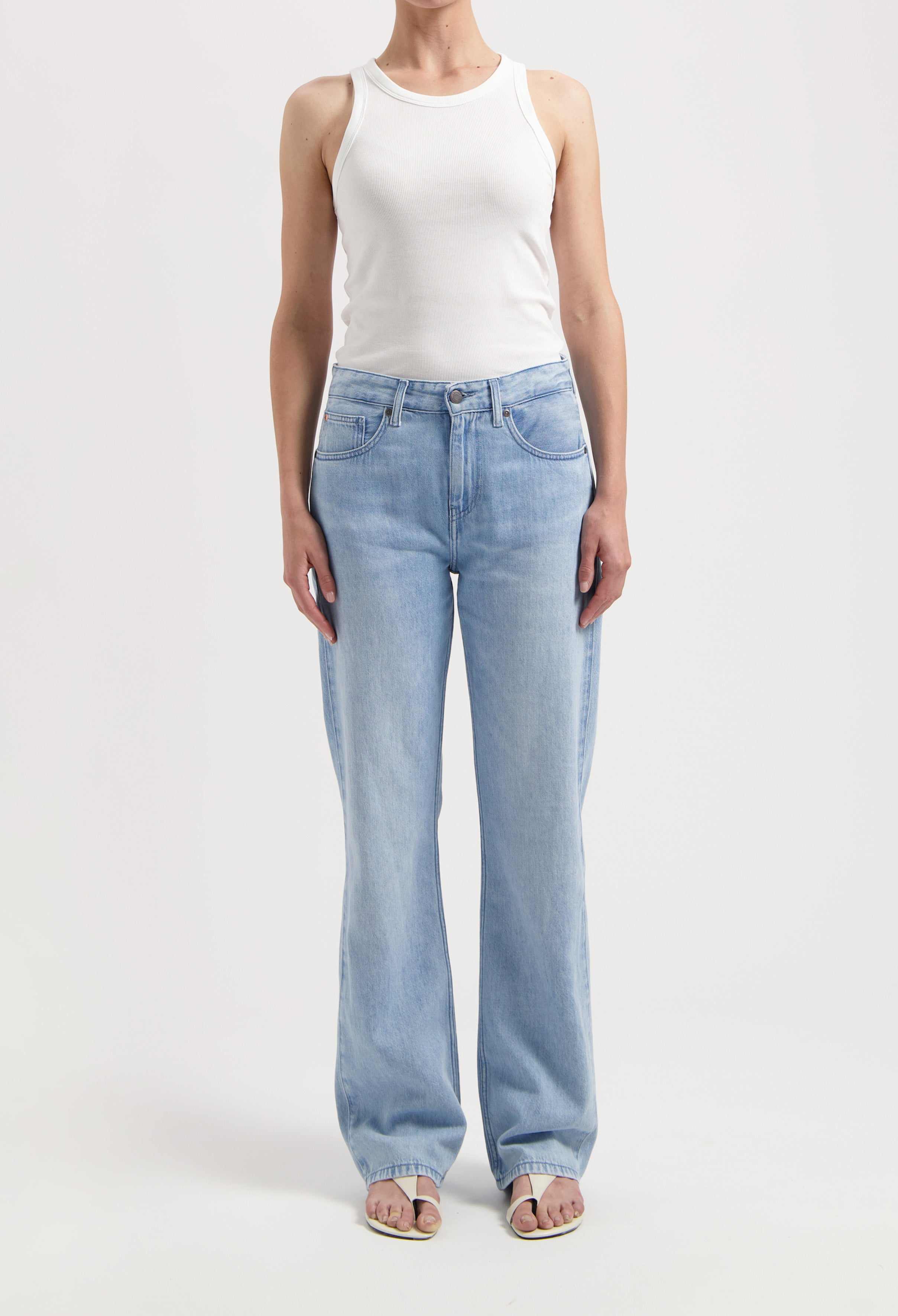 Model wearing light blue wide-leg jeans styled with a white sleeveless ribbed tank top and open-toe sandals on a plain background.