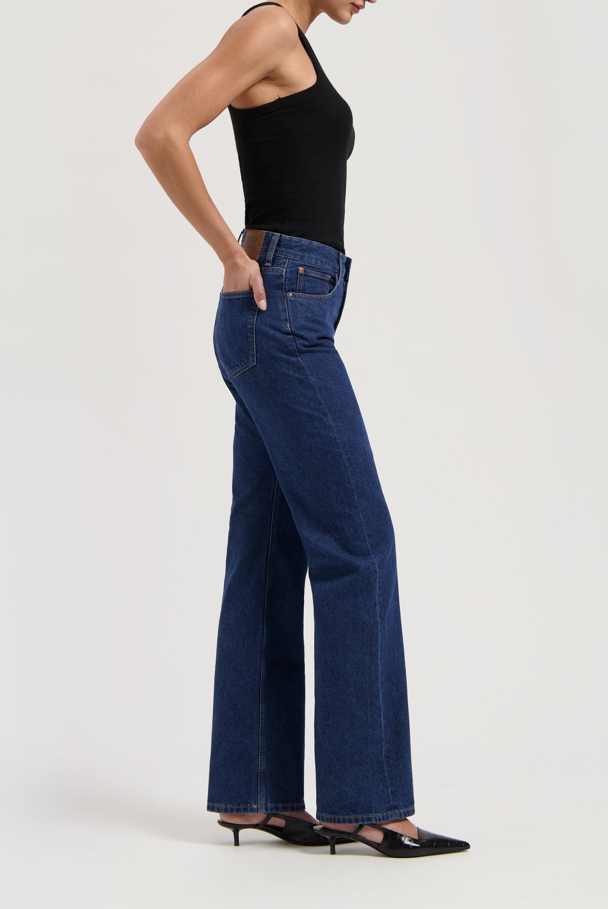 Model wearing high-waisted indigo denim jeans with a black sleeveless top and black heeled shoes on a neutral background.