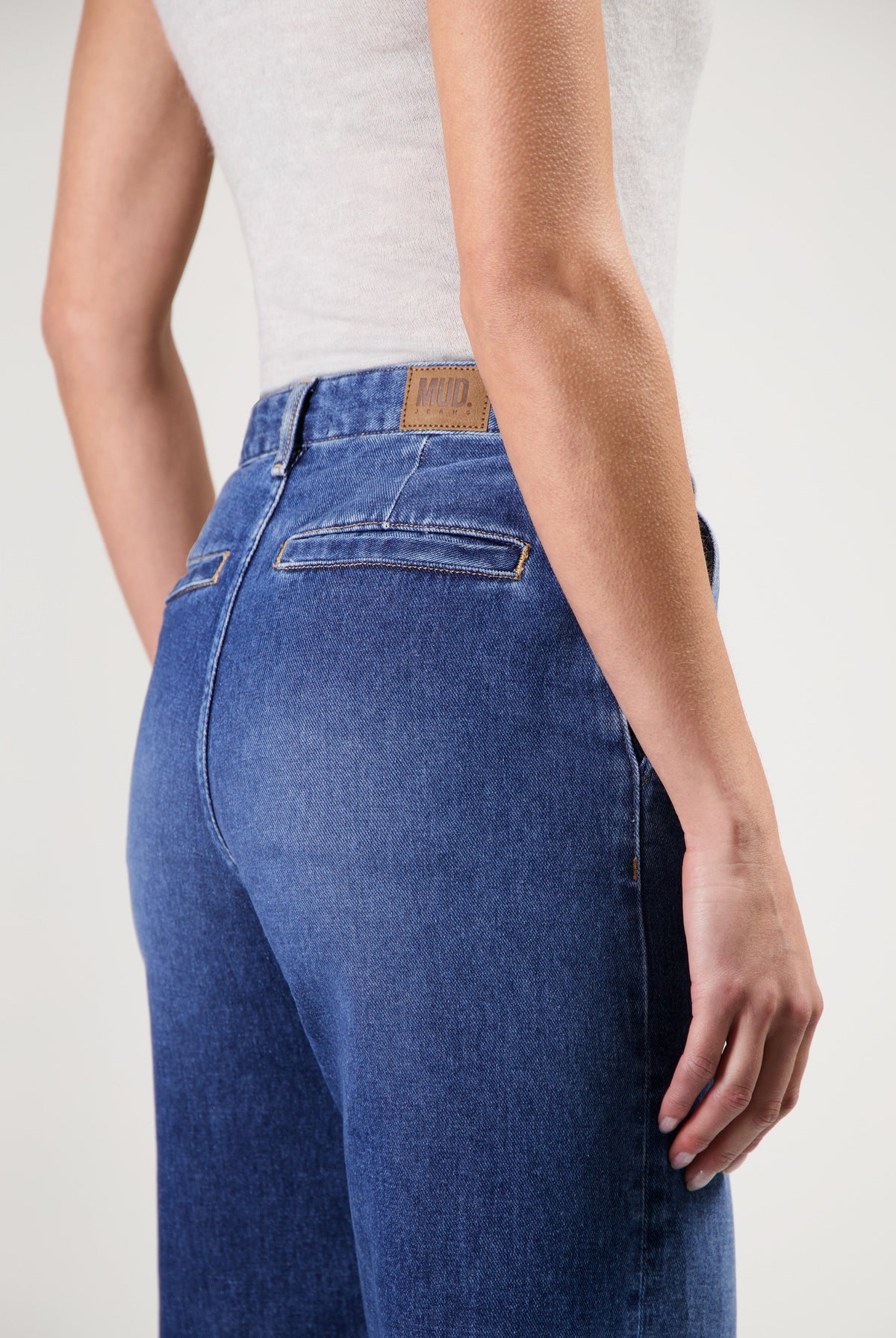 High-waisted sustainable MUD Jeans in blue, showcasing back details with brown label, worn with a light gray tank top.