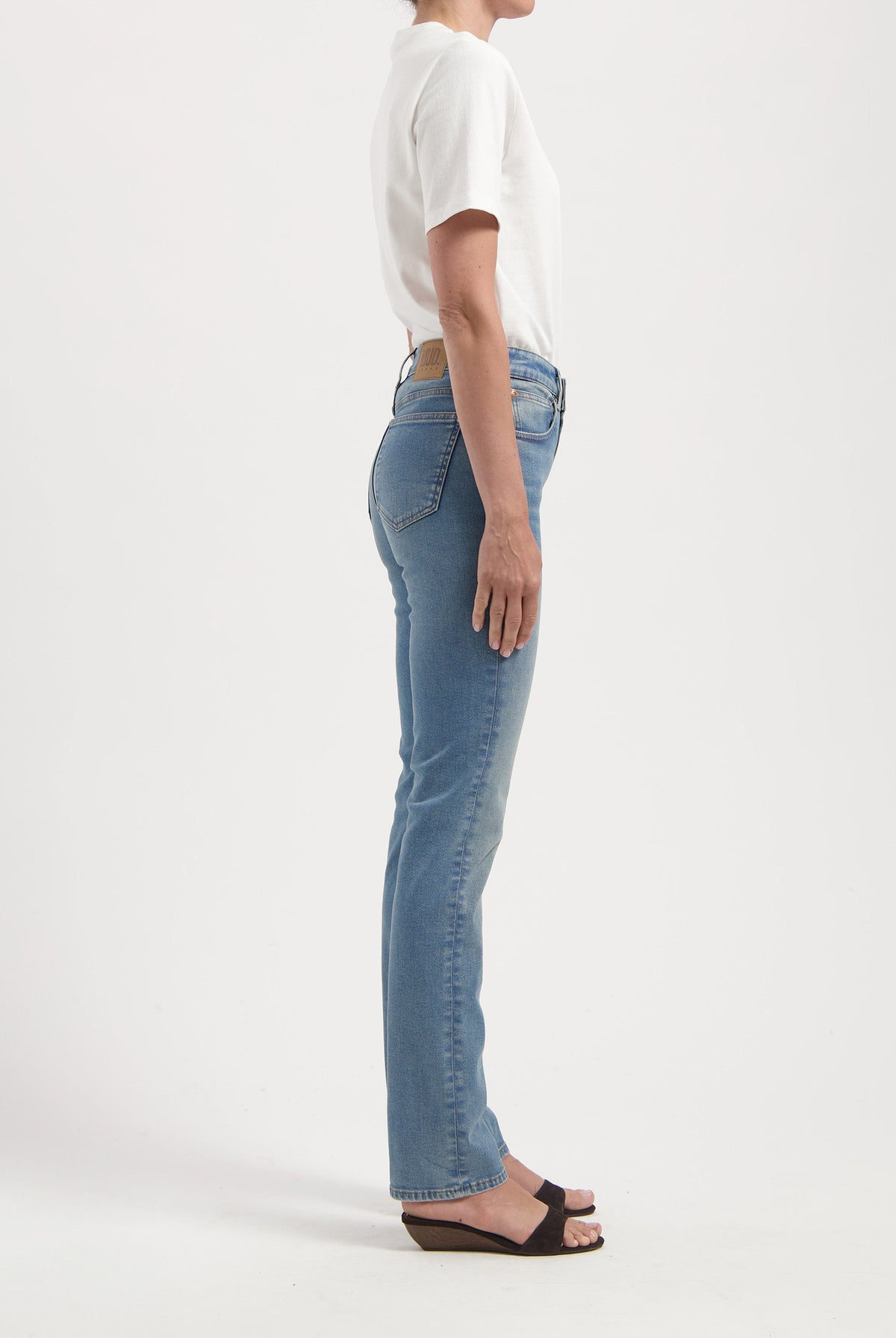 A side view of a person wearing light blue straight-leg jeans and a white short-sleeve shirt, paired with black wedge sandals.