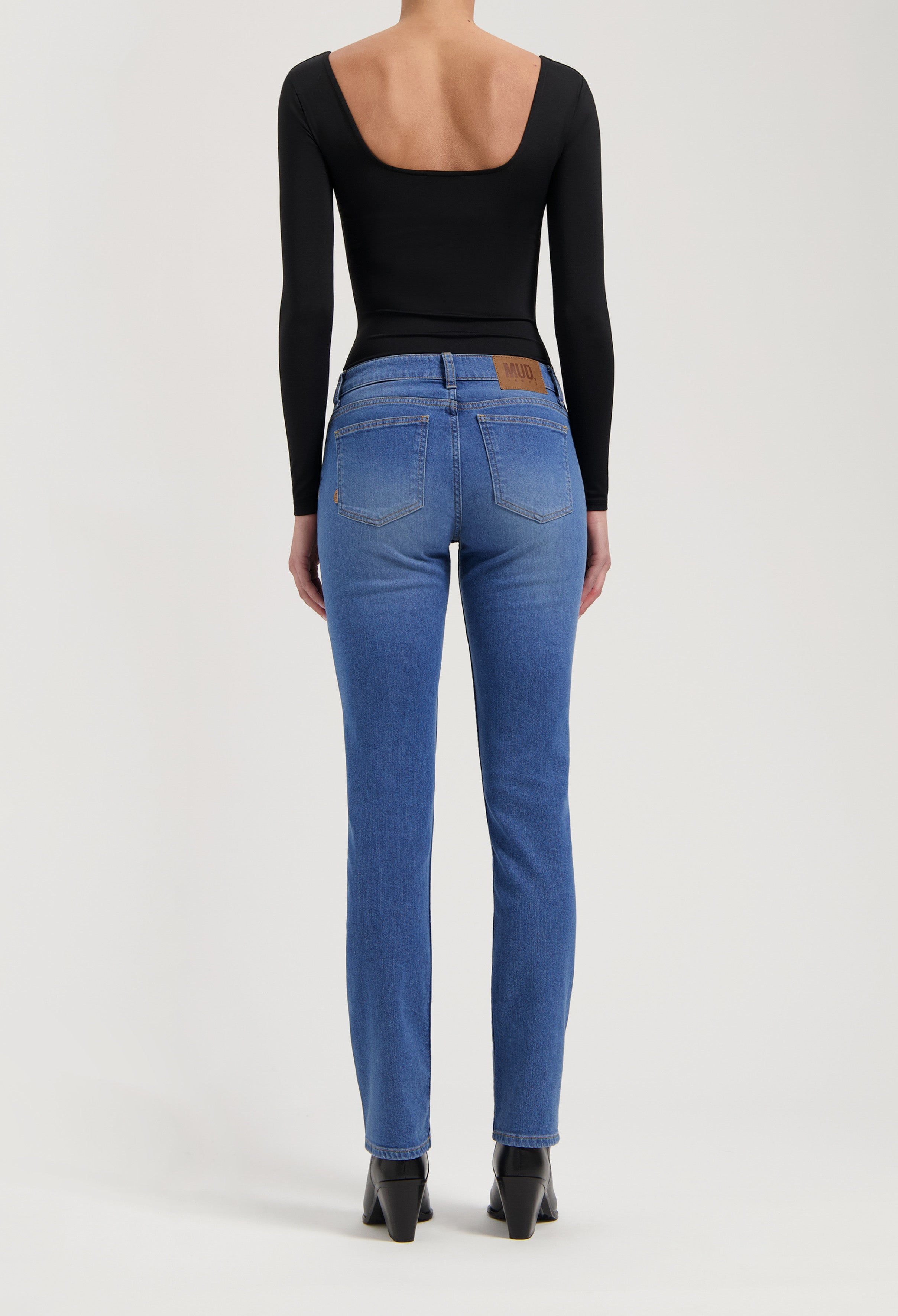 Rear view of MUD organic cotton jeans in blue, paired with a black scoop-back long-sleeve top and black ankle boots.