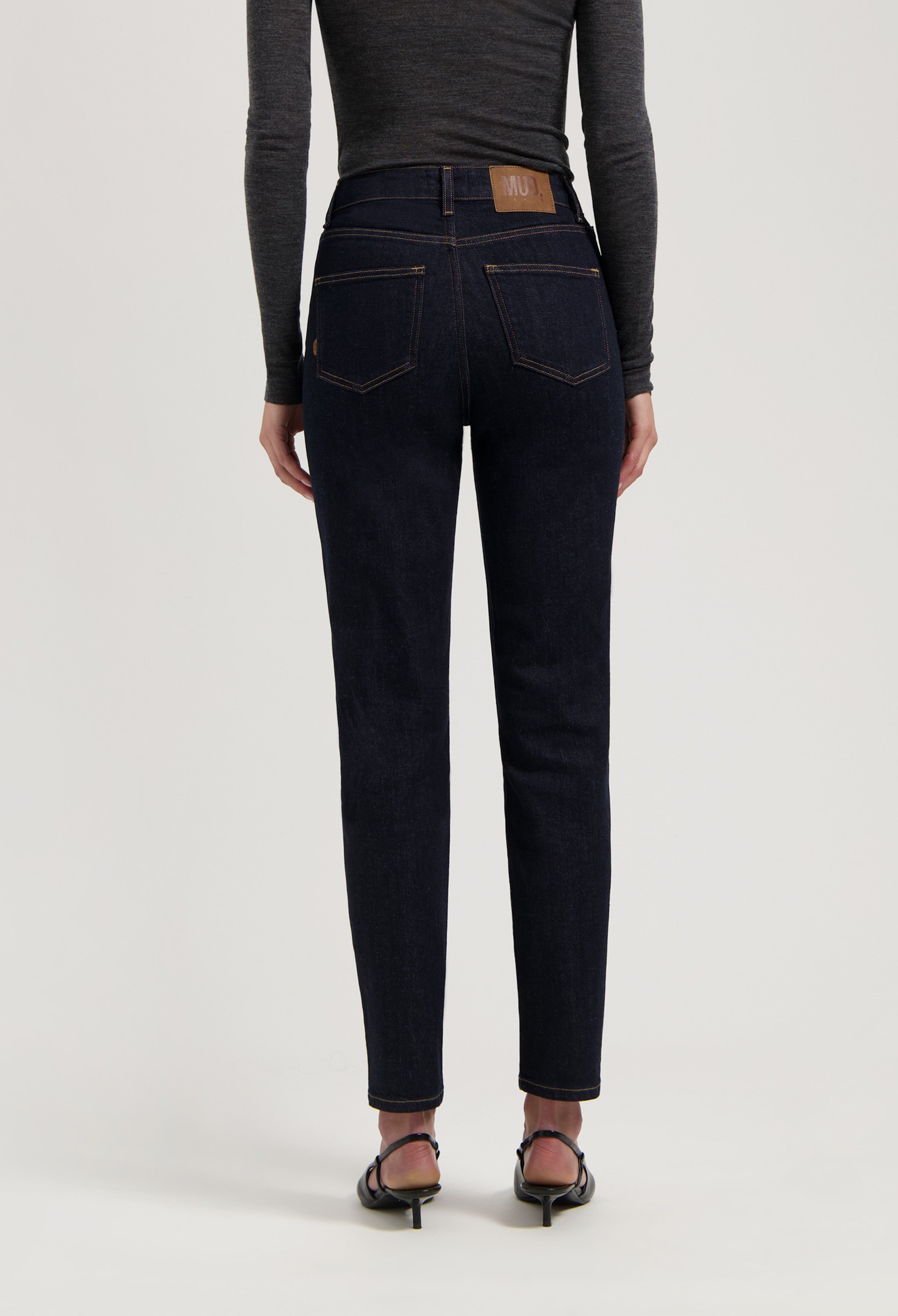 Dark indigo slim-fit jeans with classic stitching, worn with gray long-sleeve top and black-heeled sandals, rear view.