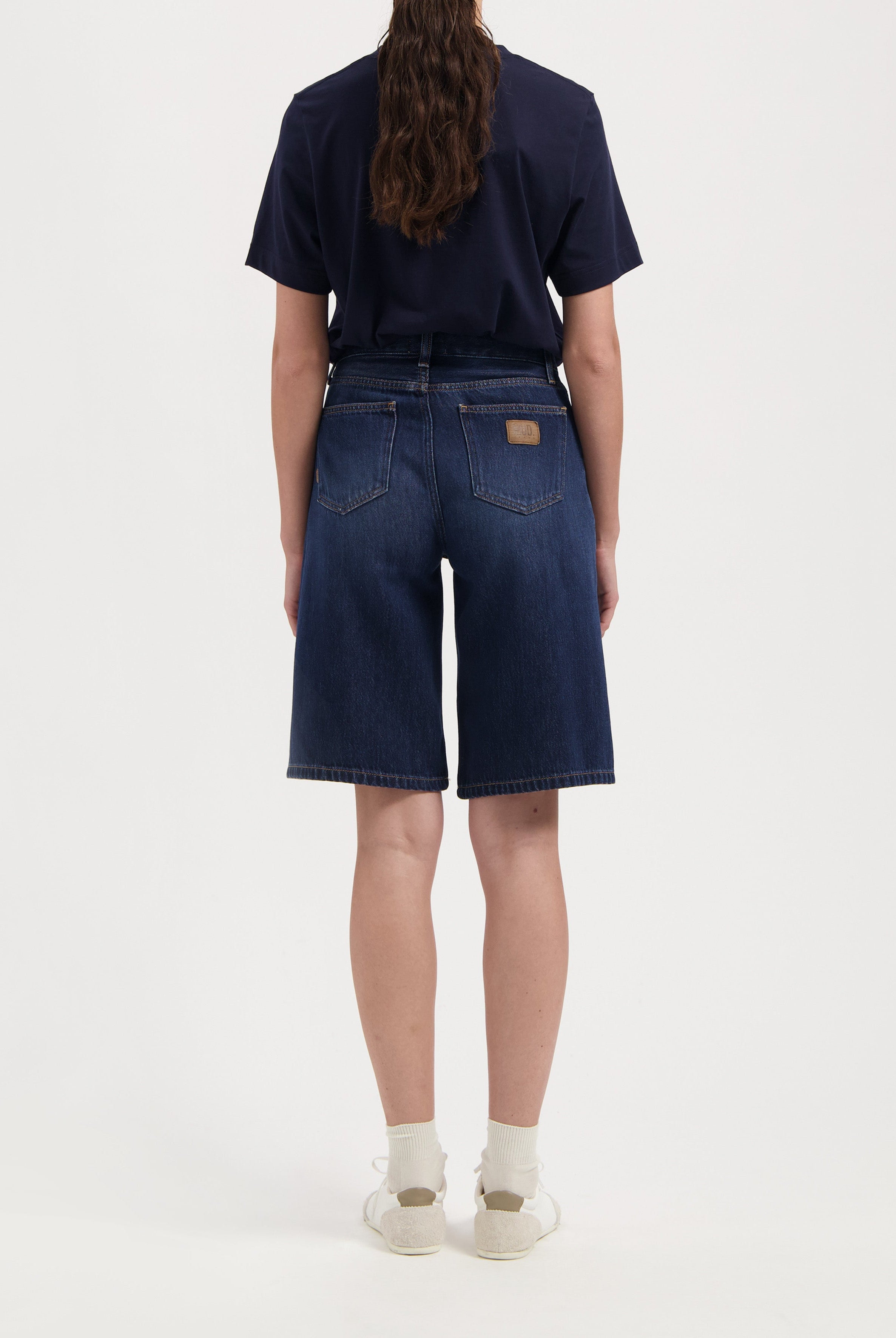 Back view of dark blue denim shorts paired with a black tee, showcasing relaxed fit and classic pocket design with white sneakers.