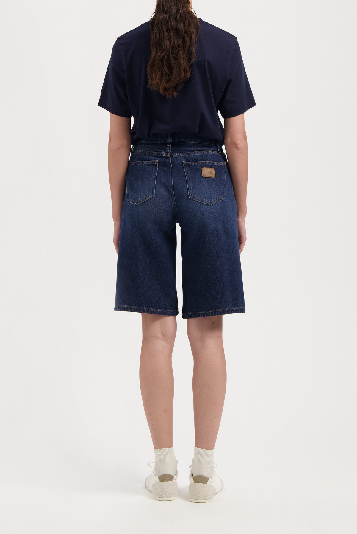 Back view of dark blue denim shorts paired with a black tee, showcasing relaxed fit and classic pocket design with white sneakers.
