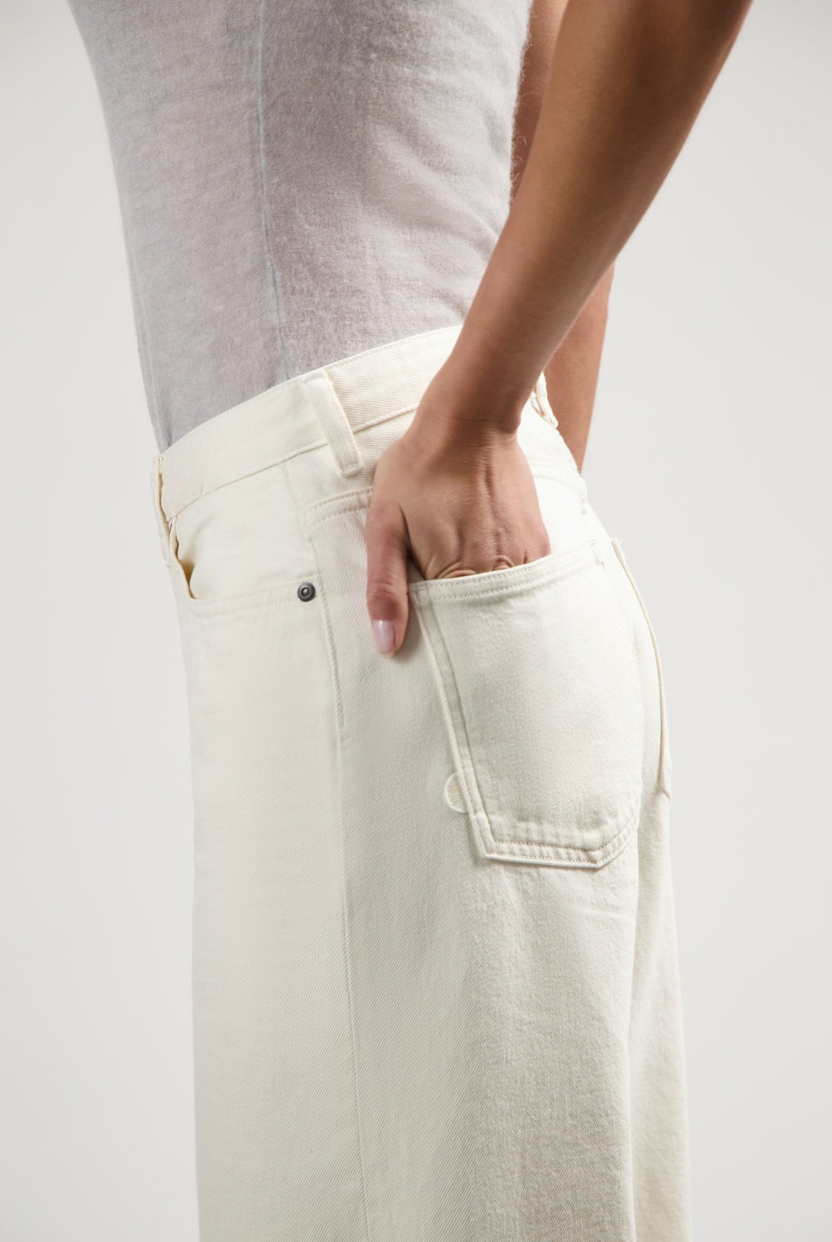 Person wearing off-white high-waisted jeans with a fitted gray sleeveless top, hand in back pocket, side view.