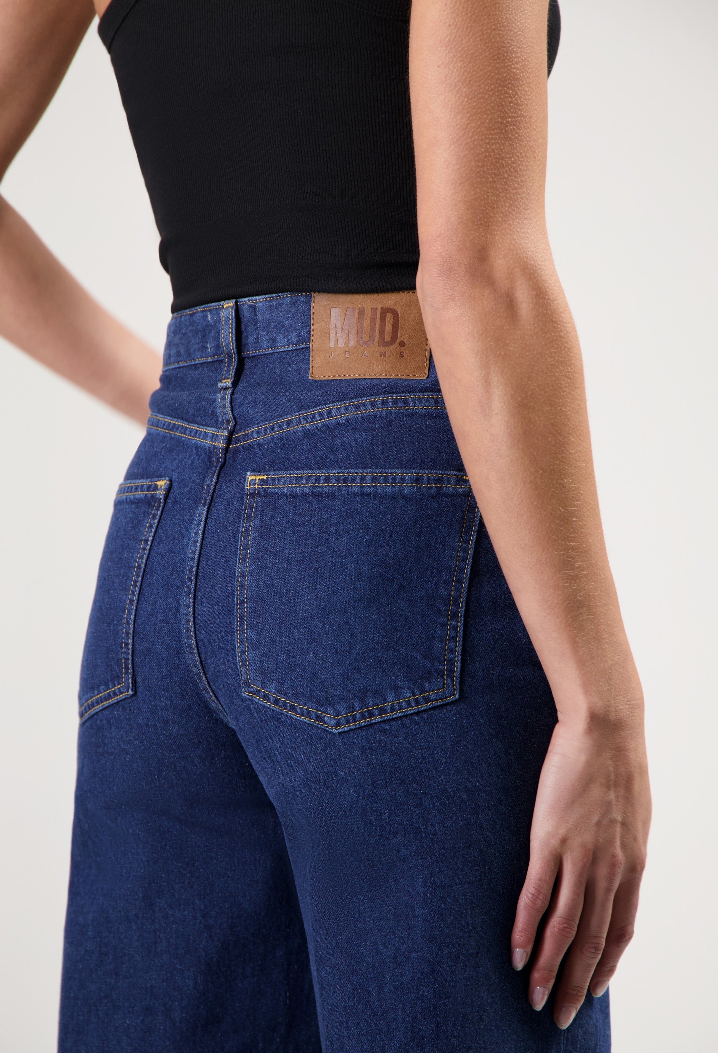Close-up of indigo blue MUD Jeans made from organic cotton, showing back pockets and branding, paired with a black tank top.