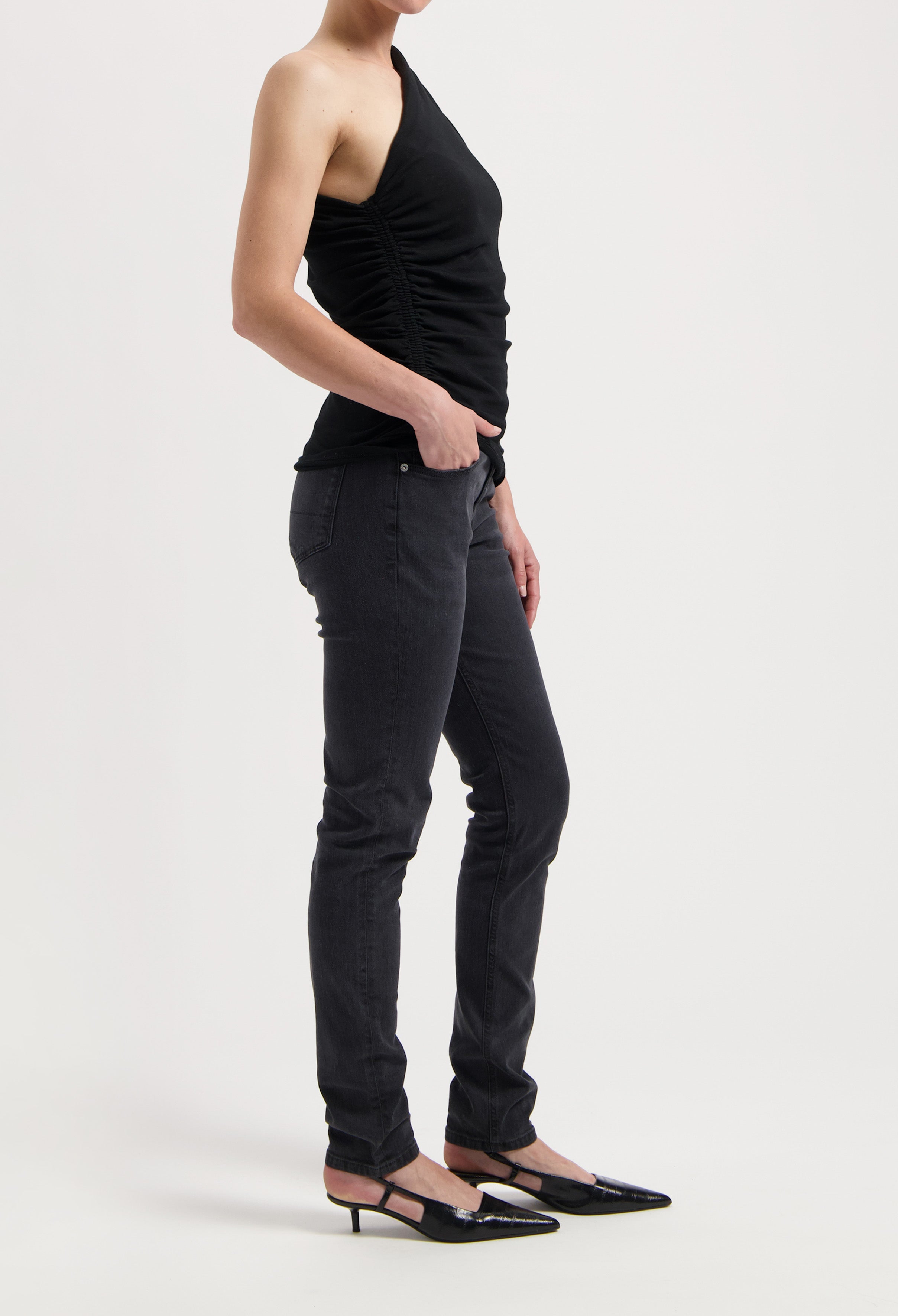 One-shoulder black top styled with charcoal slim-fit jeans and black pointed-toe heels on white background.