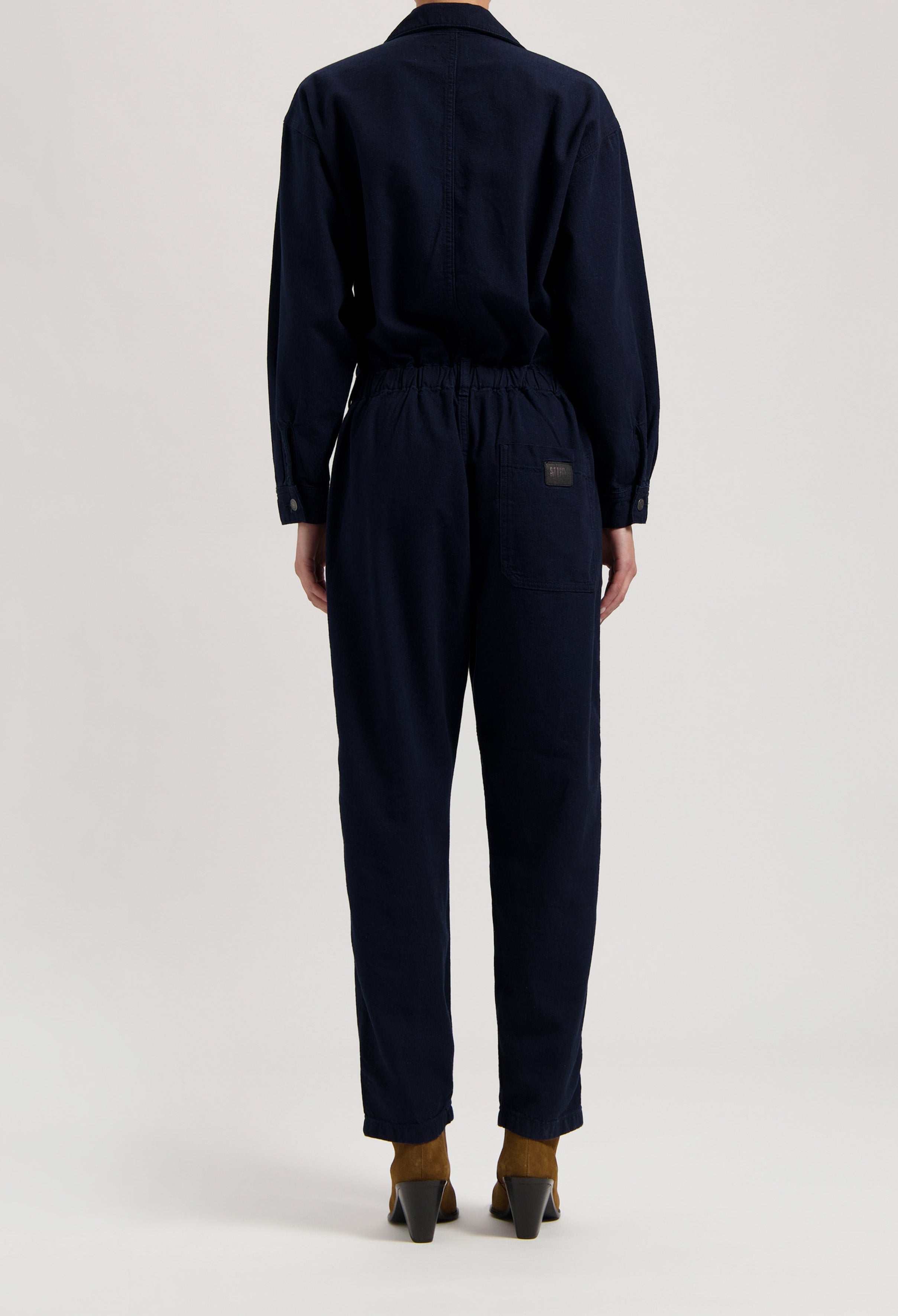 Rear view of a navy blue cotton jumpsuit with an elastic waistband, paired with brown ankle boots and black buttons.