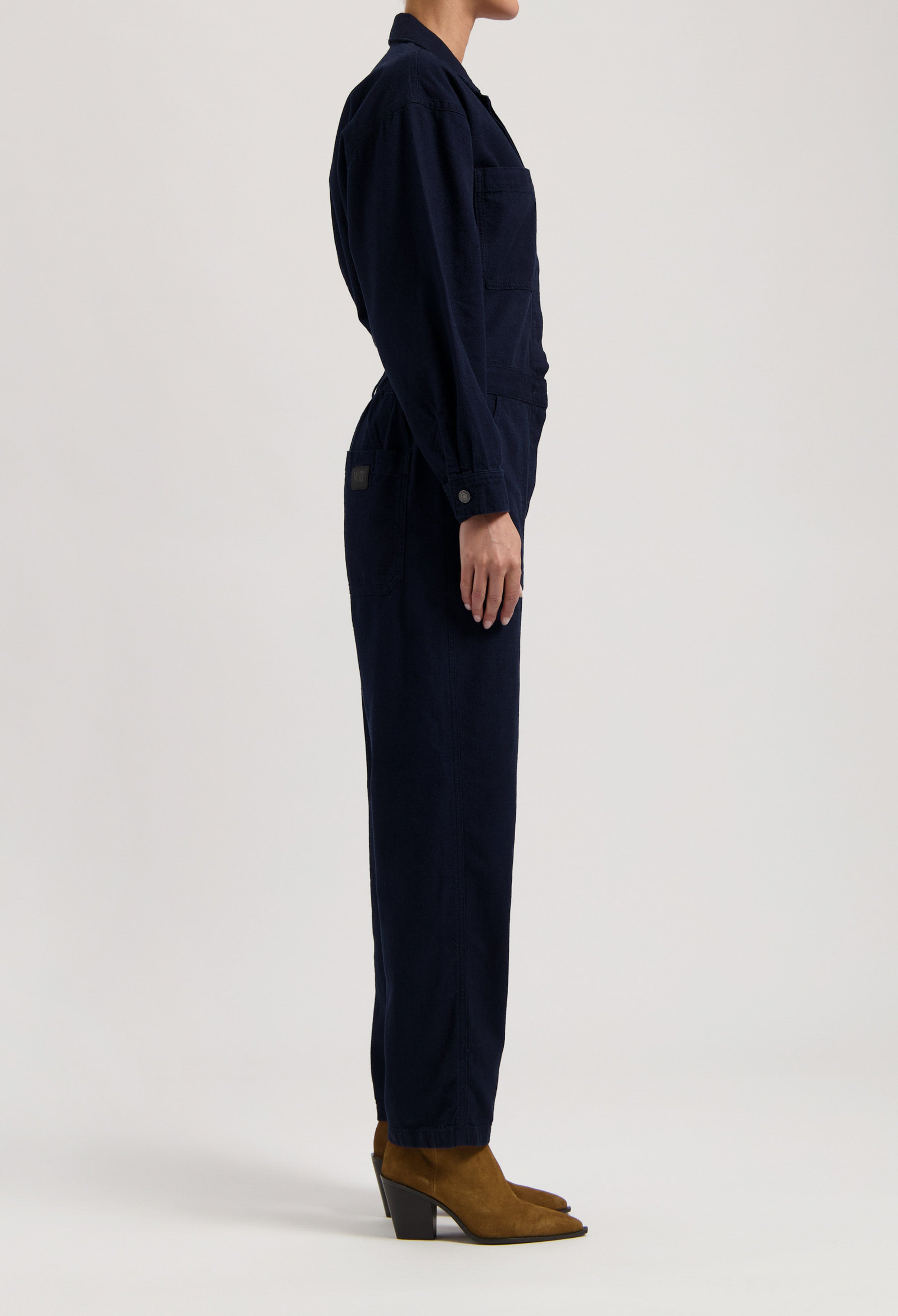 Navy blue cotton utility jumpsuit with long sleeves, side pockets, and brown suede ankle boots, showcased in side profile.