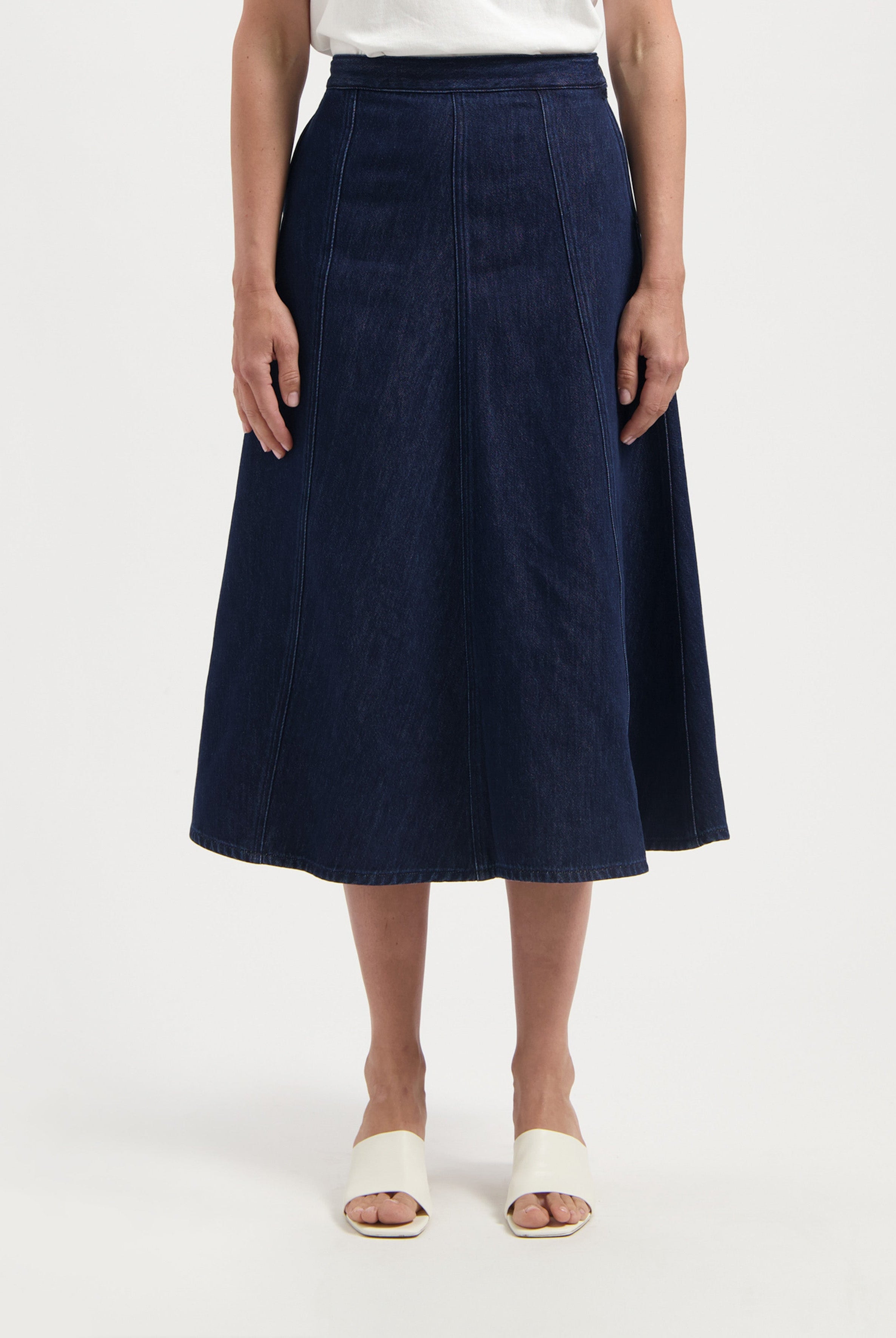 Dark blue, A-line denim skirt worn with a white top, showcasing a classic midi length and vertical seam detailing.