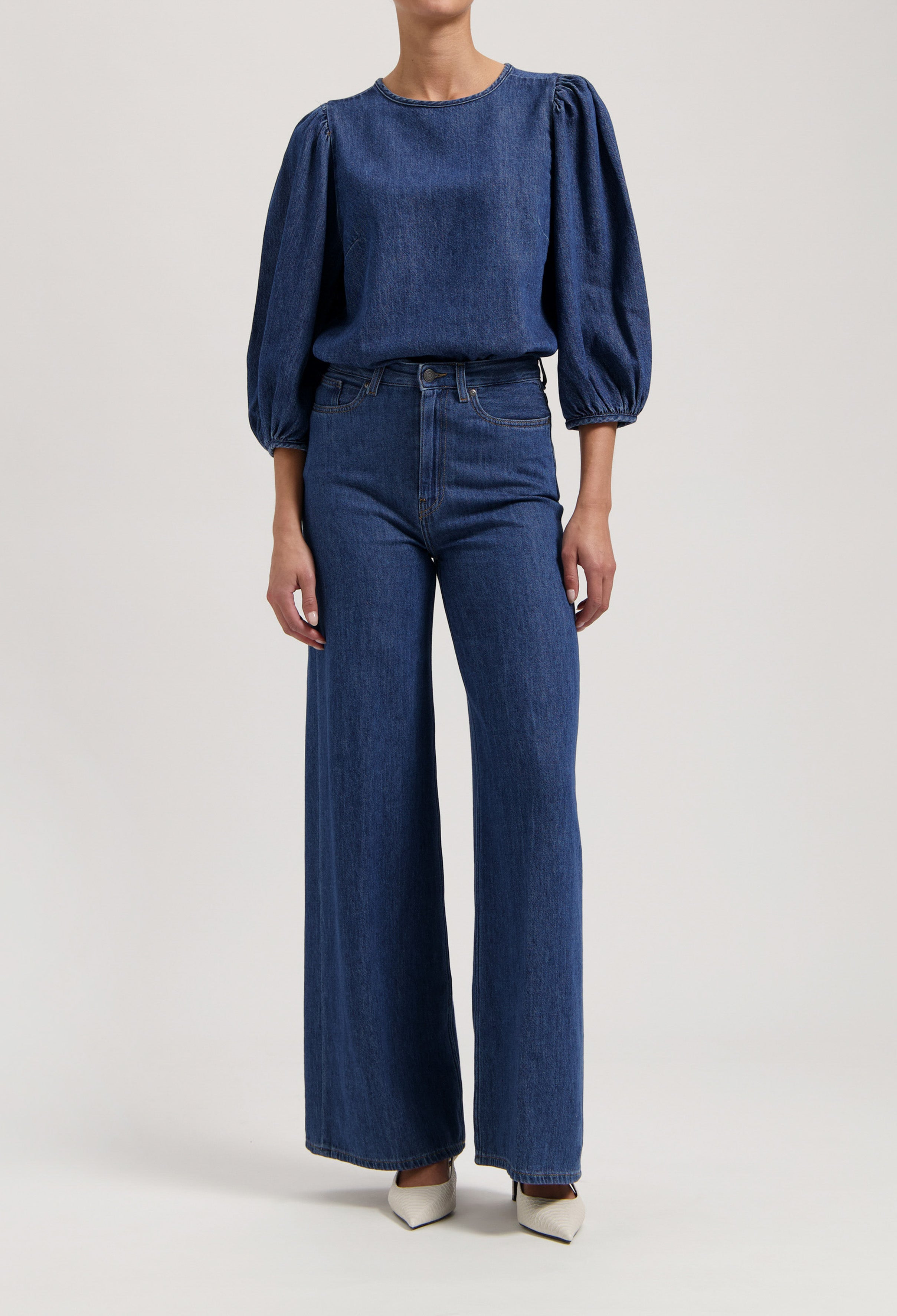 Woman wearing dark blue denim top with puff sleeves and matching high-waisted wide-leg jeans, paired with white pointed heels.
