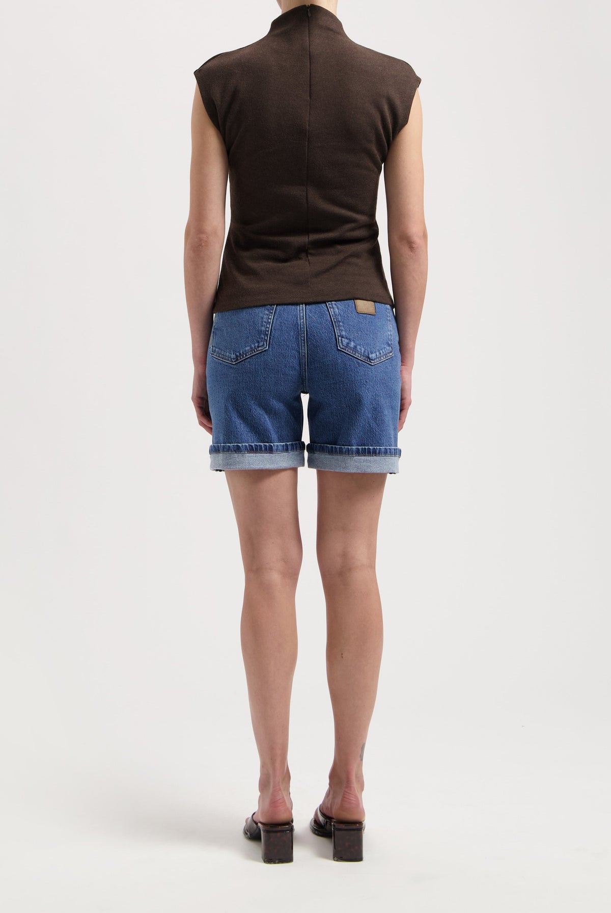 Woman wearing blue denim shorts and sleeveless dark brown top, rear view, paired with black block-heeled sandals.
