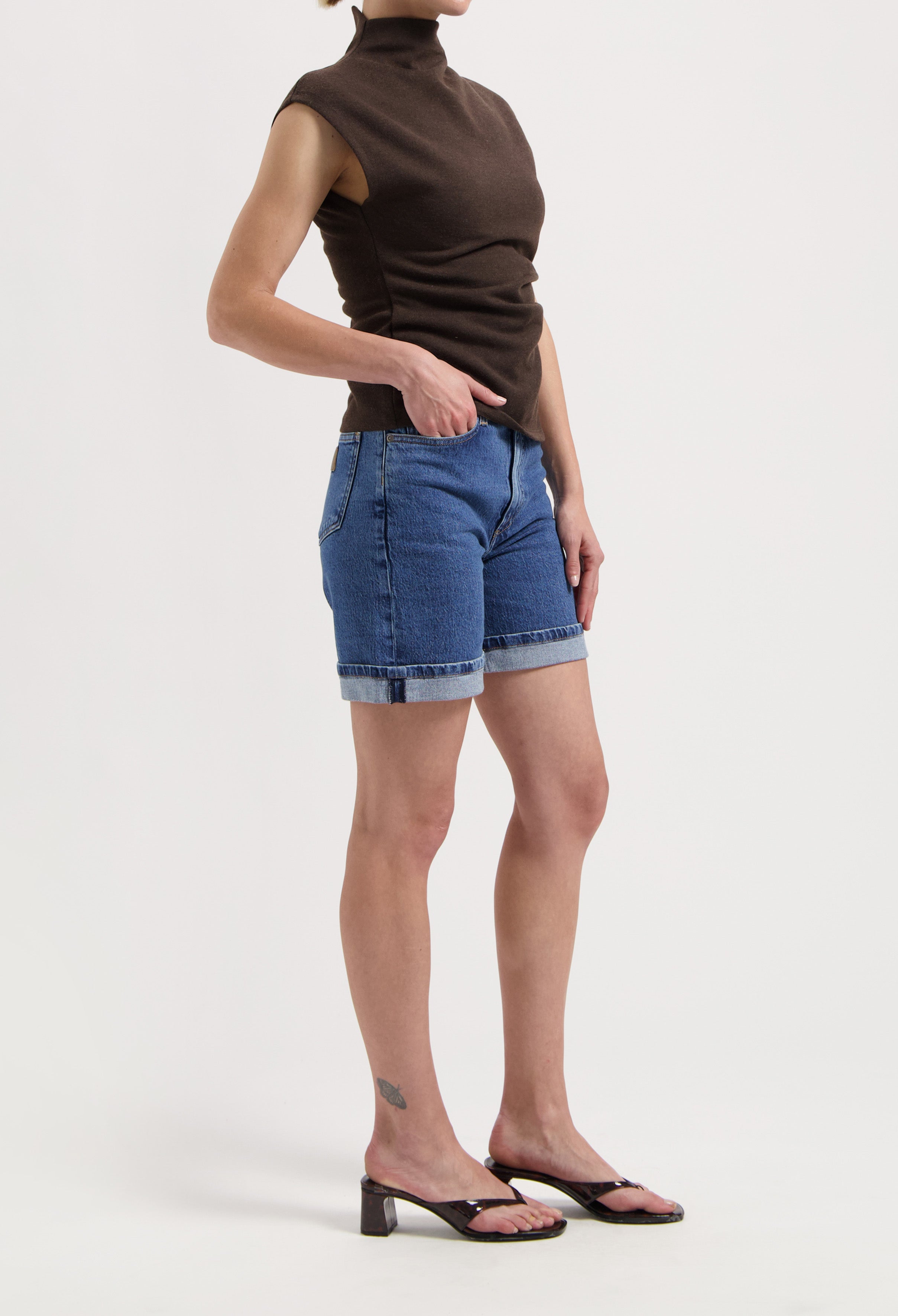 Woman wearing denim shorts with rolled cuffs paired with a sleeveless dark brown high-neck top and black heeled sandals.
