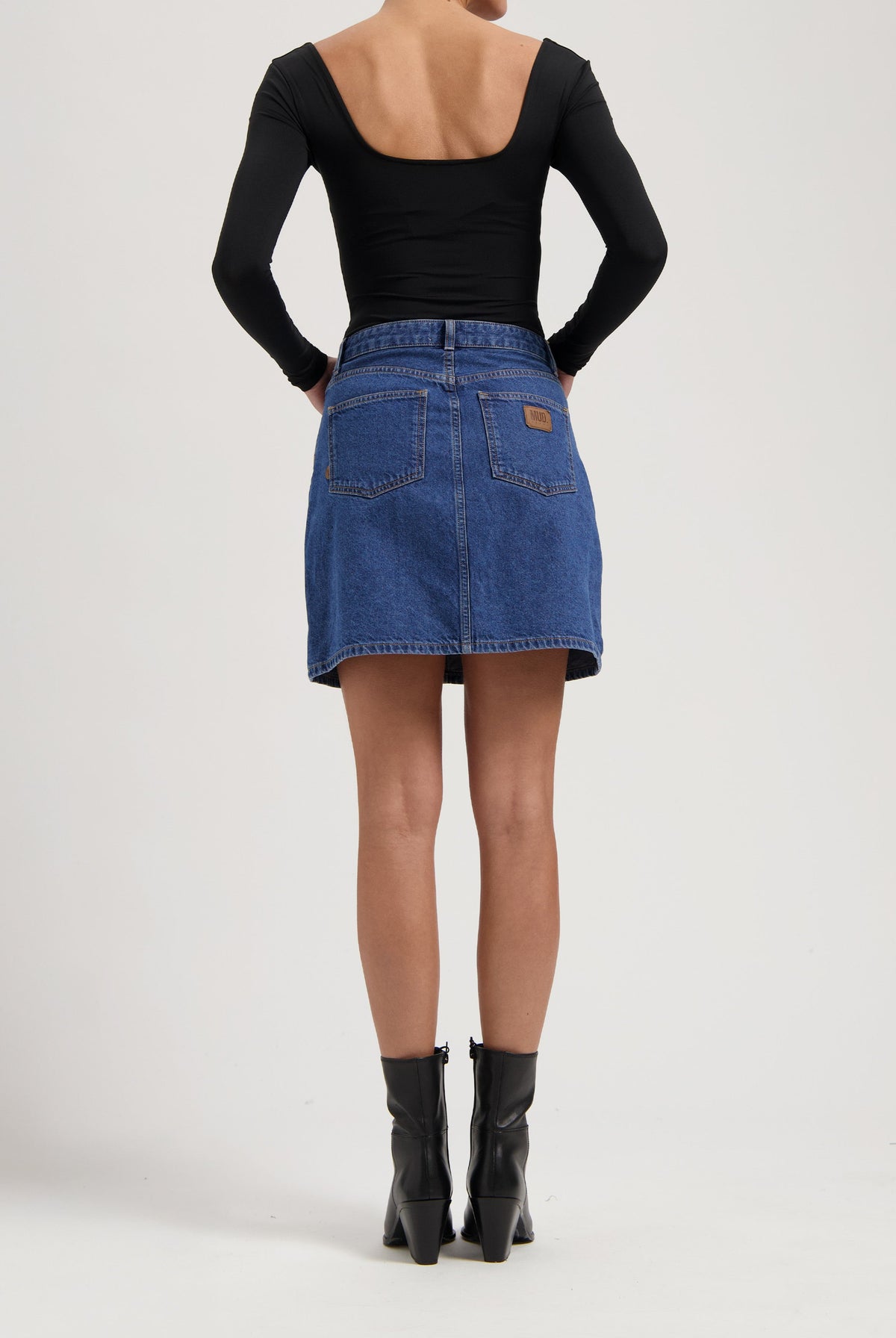 Back view of a woman in a black, long-sleeve top and a blue denim skirt, paired with sleek, black ankle boots.