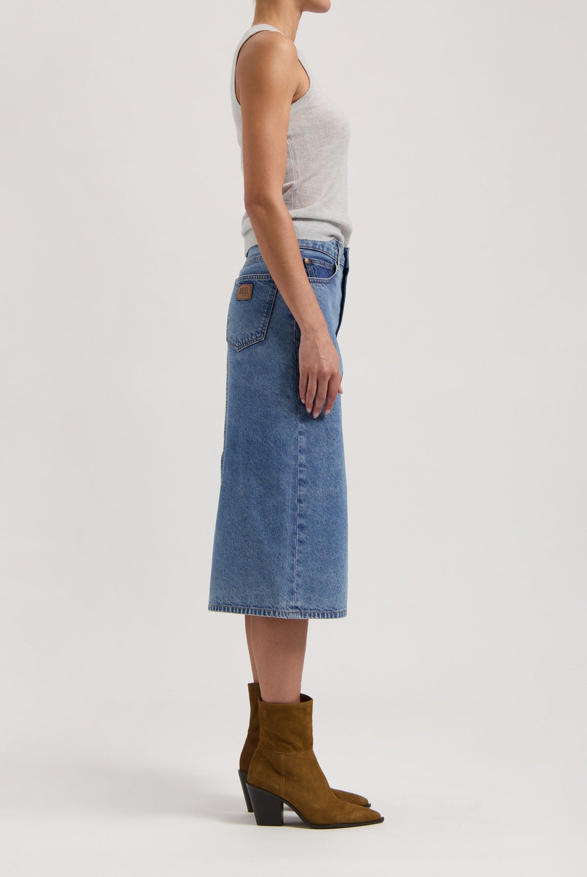 Side view of a woman in a sleeveless gray top, wearing a blue denim midi skirt paired with brown suede ankle boots.