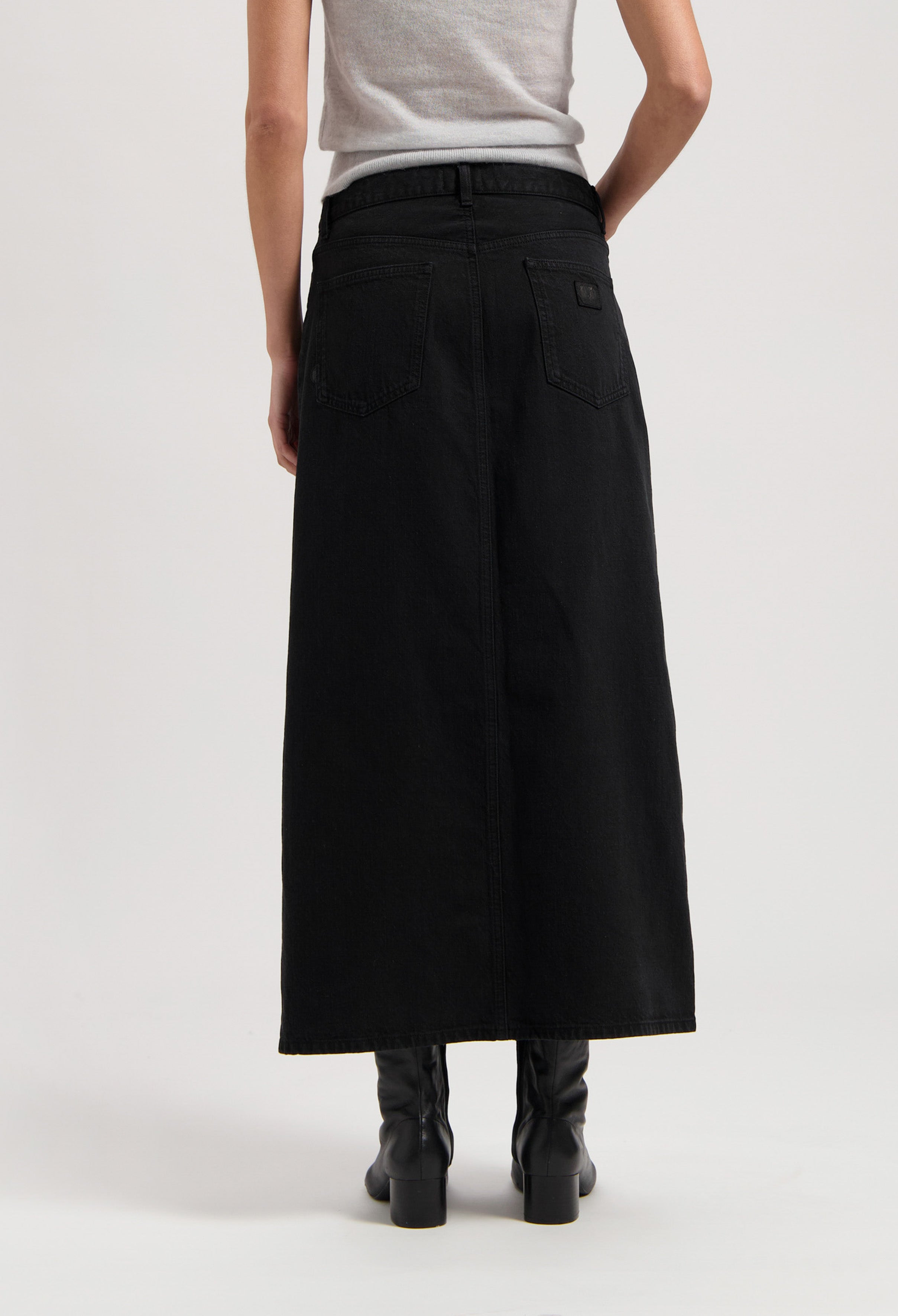 Black denim maxi skirt with back pockets worn by a model in a gray sleeveless top and black boots, showcasing back view.