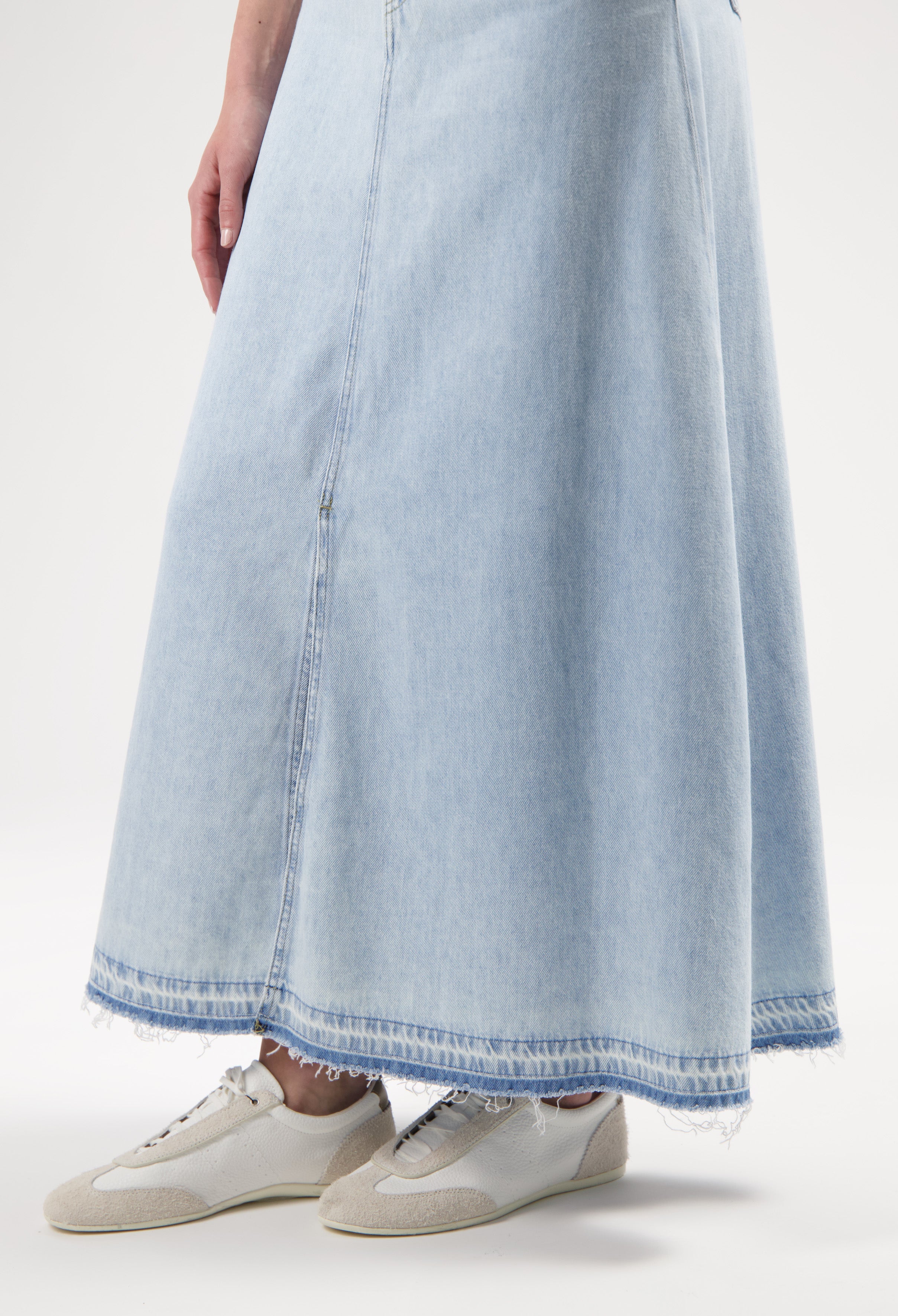 Light blue denim maxi skirt with frayed hem, paired with white and beige canvas sneakers in a minimalist style.