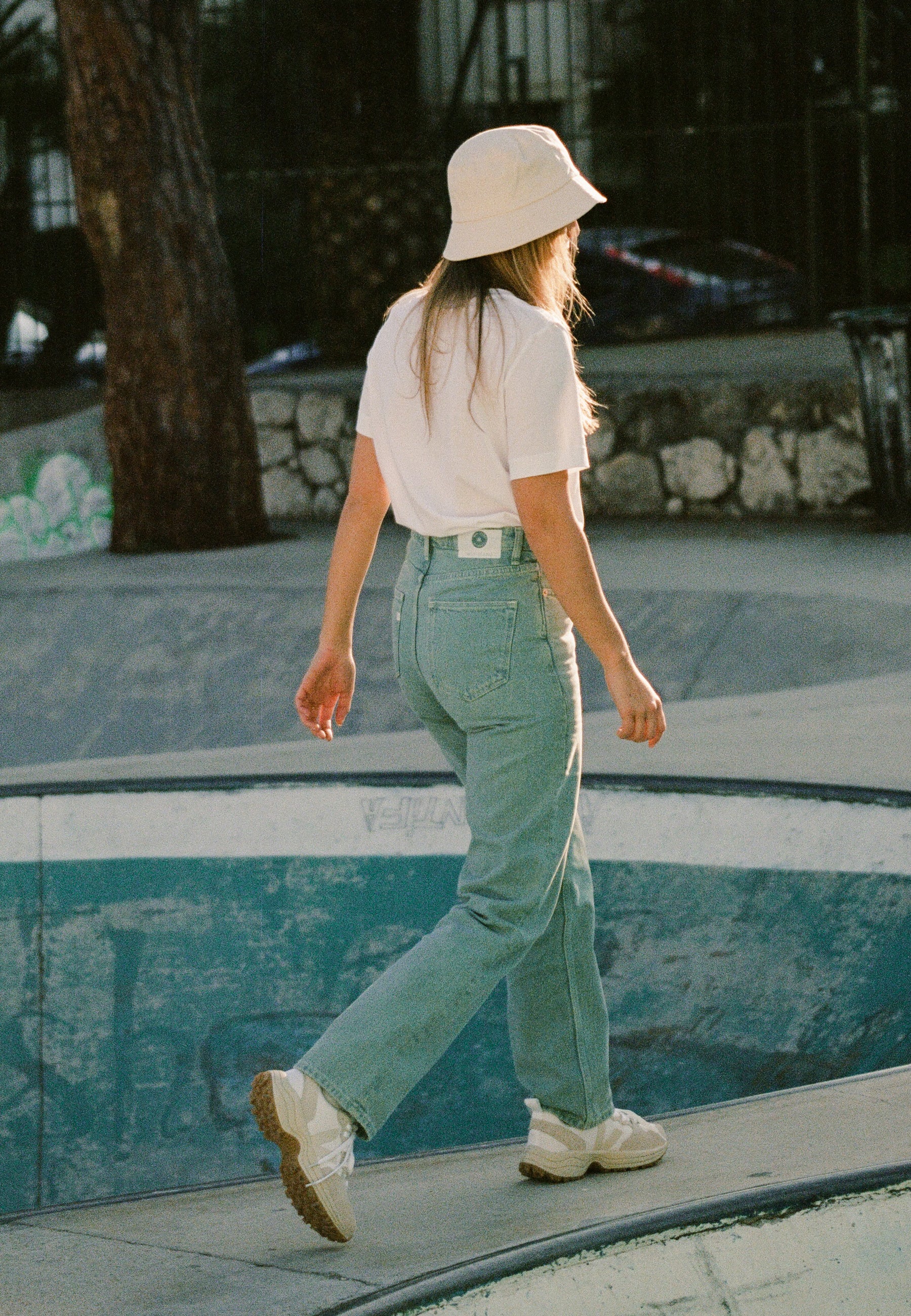 Woman walking at a skate park wearing a beige bucket hat, white t-shirt, light blue jeans, and white sneakers.