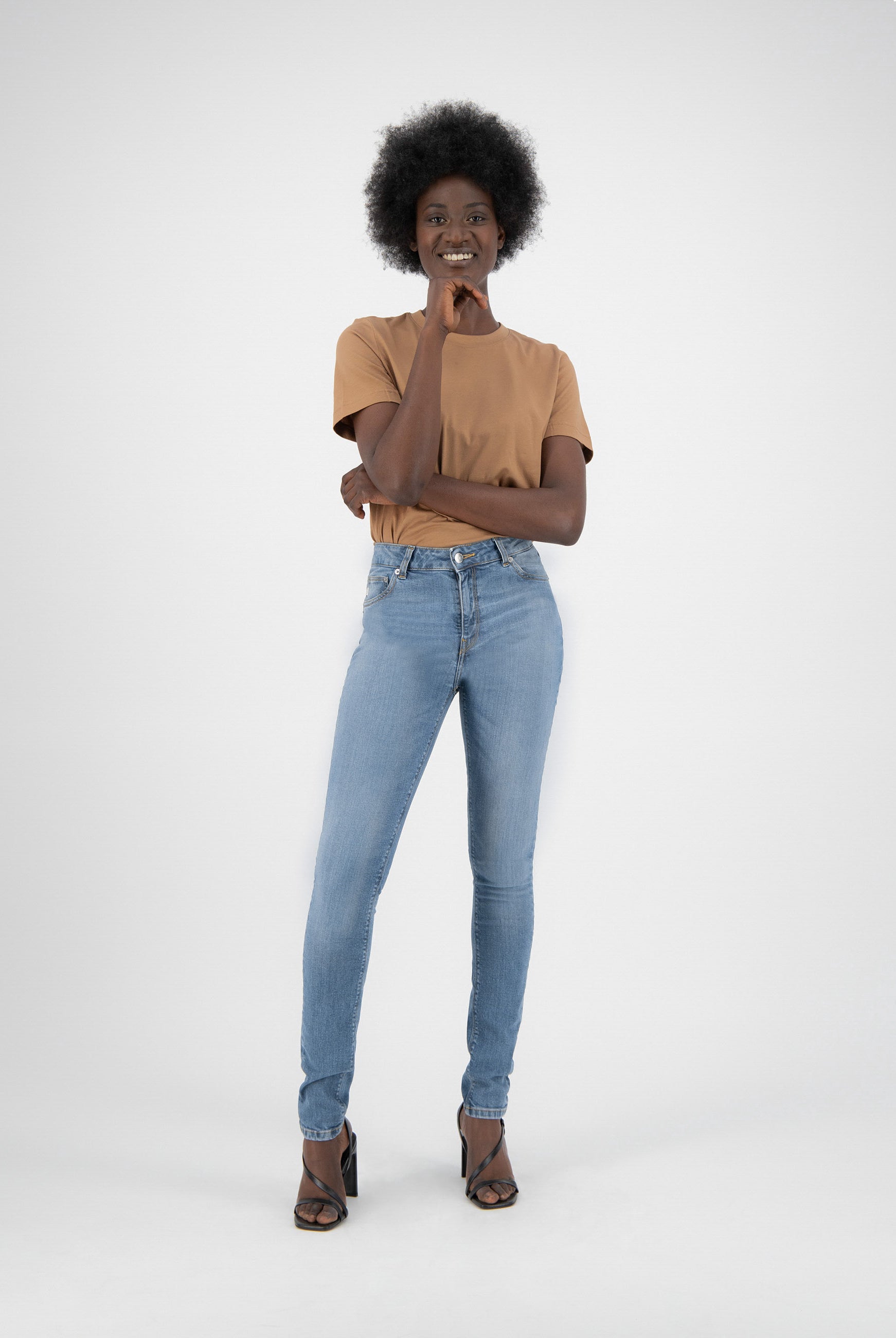 Woman modeling light blue denim jeans and tan t-shirt, wearing black heels against plain white background, smiling confidently.