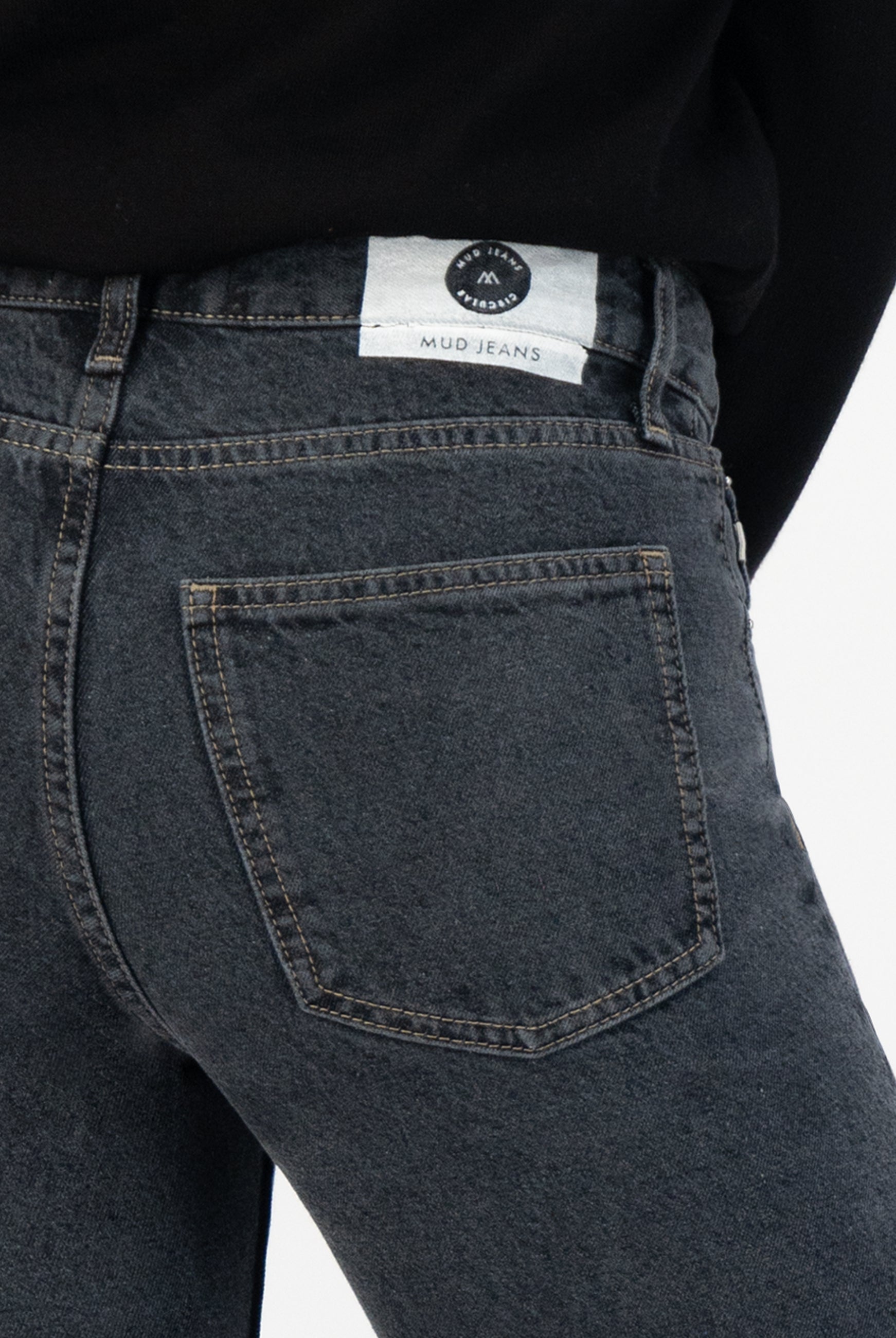 Dark gray MUD Jeans in organic cotton, highlighting close-up of back pocket stitch detail, worn with a black top.