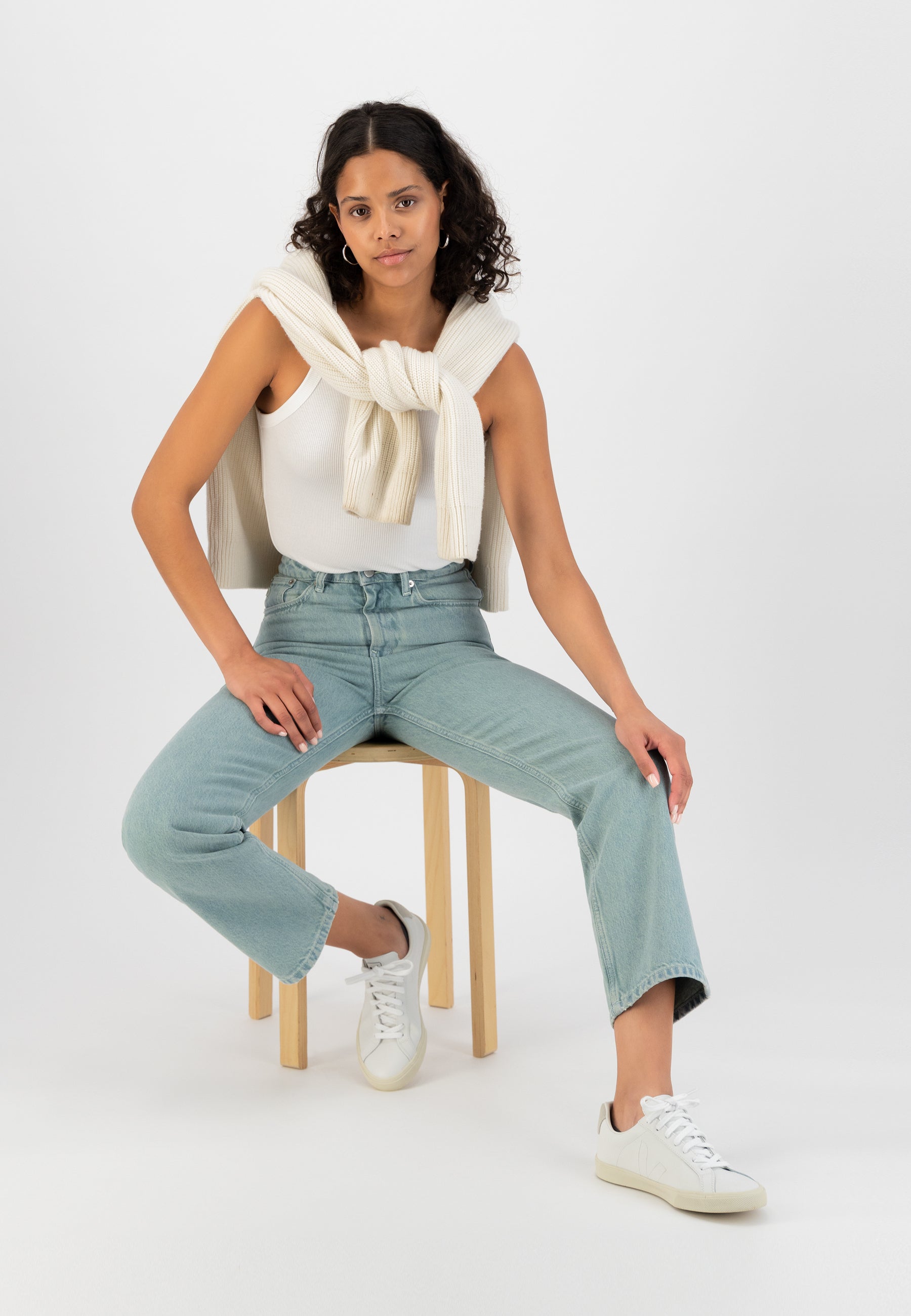 Casual fashion look featuring a white tank top, cream knit sweater, light blue jeans, and white sneakers, on a wooden stool.