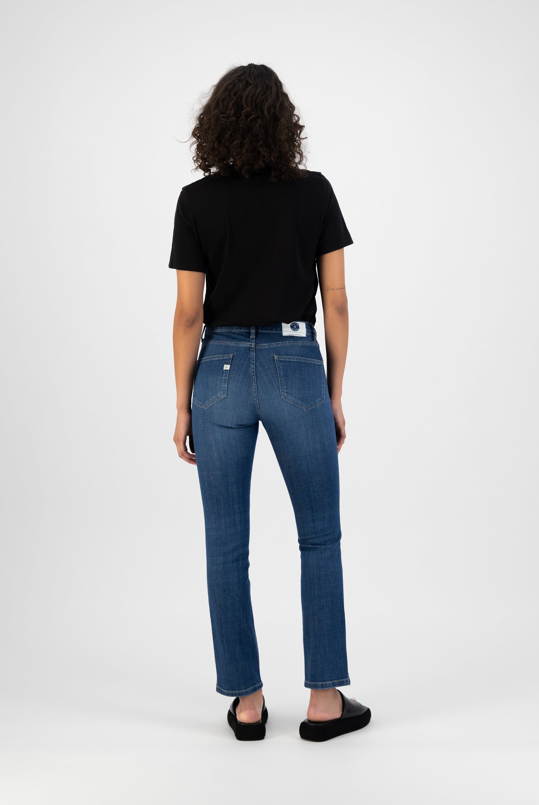 Curly-haired woman in black T-shirt and slim-fit blue jeans, showcasing back view of casual outfit with black slip-ons.