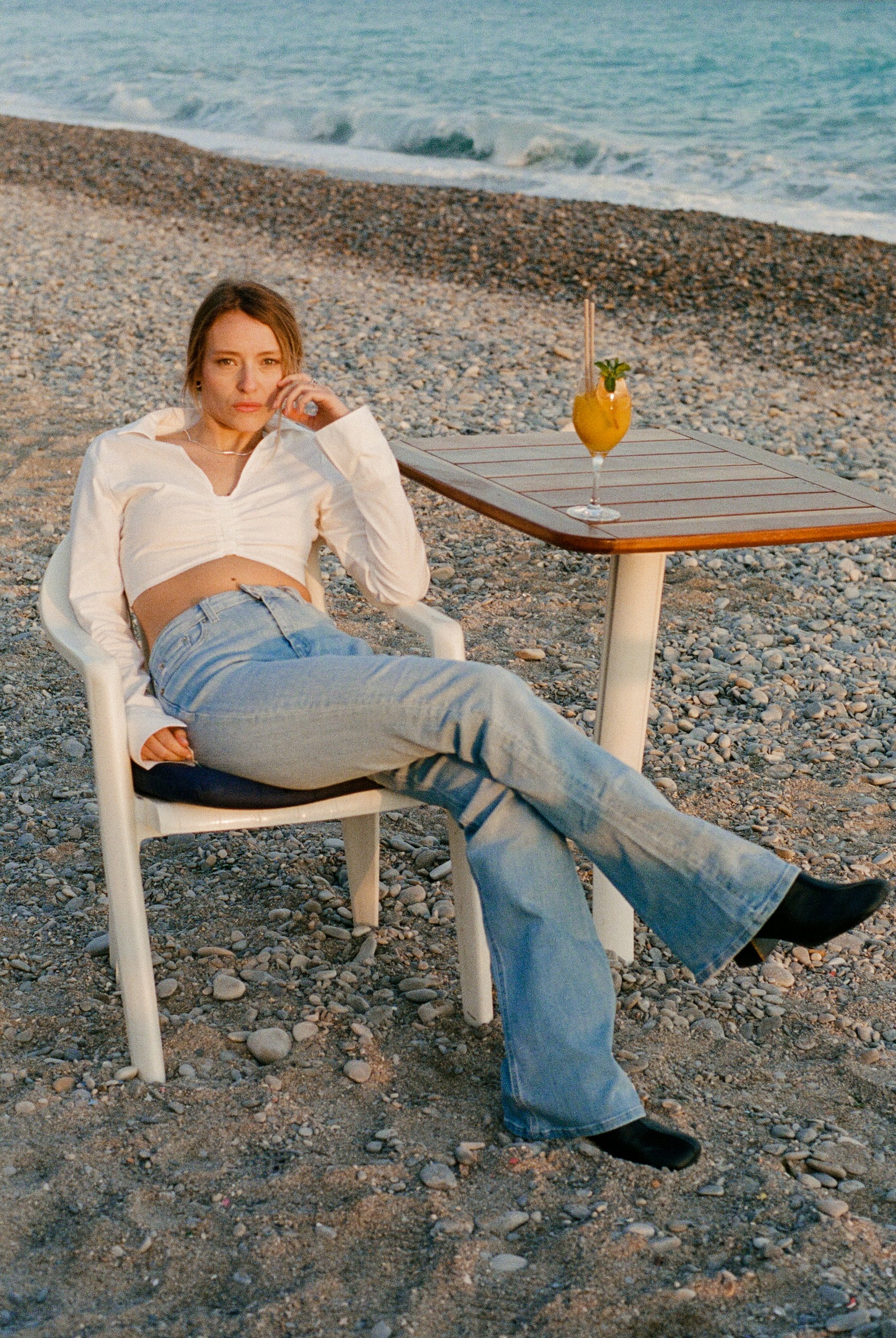 Casual beach scene with woman in light-wash jeans and white blouse, seated on a pebbled shore beside a wooden table.