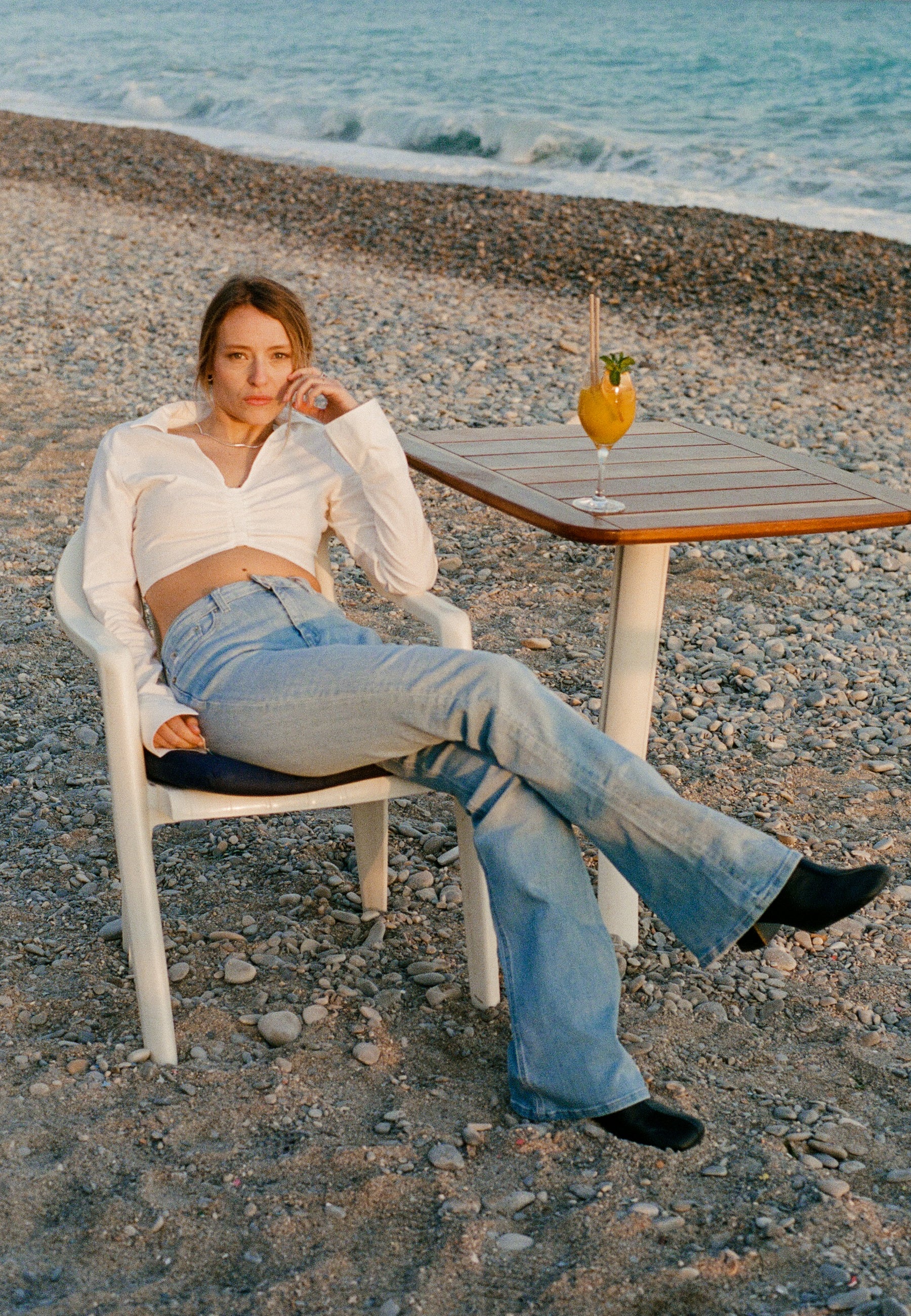 Casual beach scene with woman in light-wash jeans and white blouse, seated on a pebbled shore beside a wooden table.