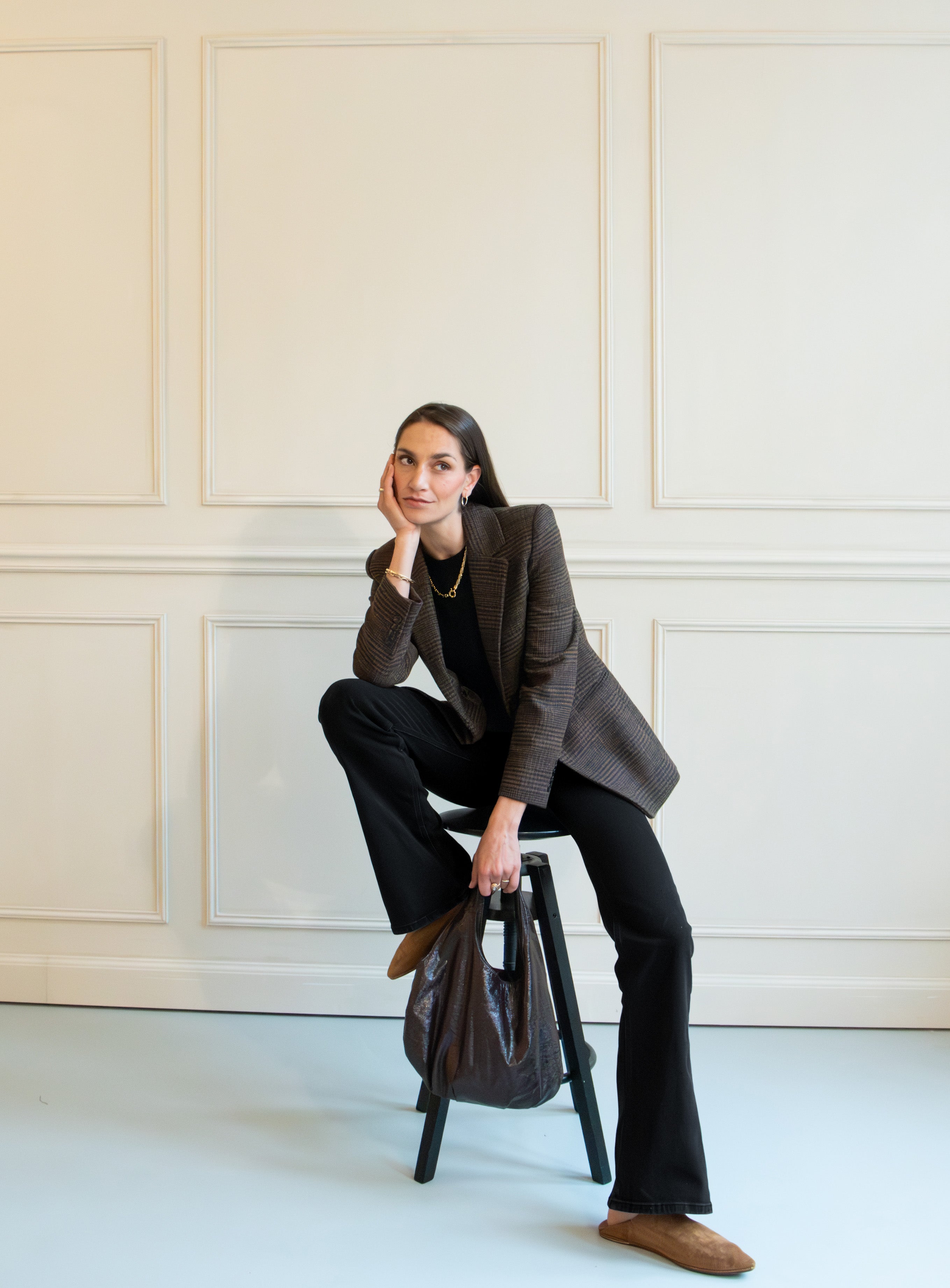 Stylish woman in a checkered blazer and black pants, seated on a stool in an elegant, cream-paneled room.