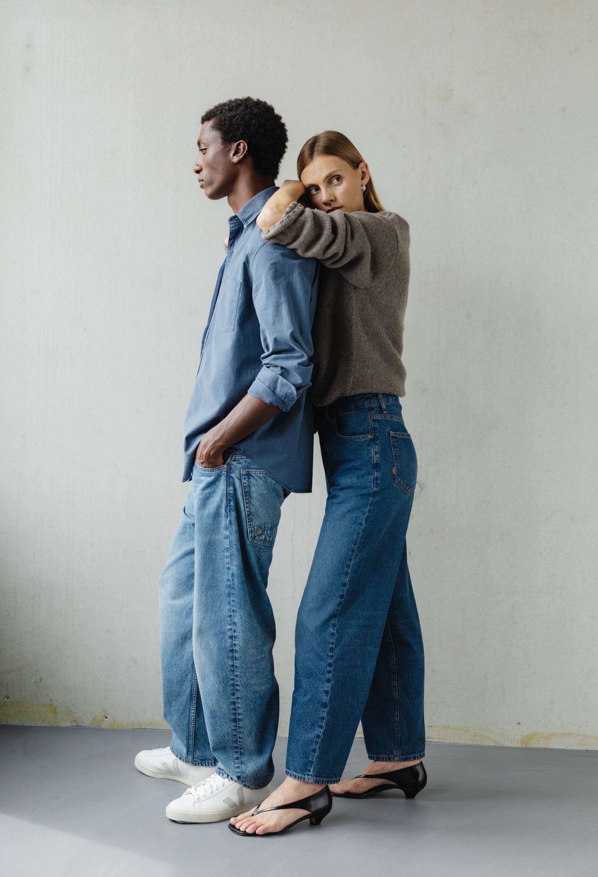 Casual fashion featuring a couple in stylish denim jeans, one in a gray sweater and black sandals, other in a blue shirt.