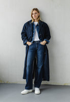 Woman wearing a stylish denim ensemble with a long coat, paired with a white t-shirt and sneakers, leaning against a wall.