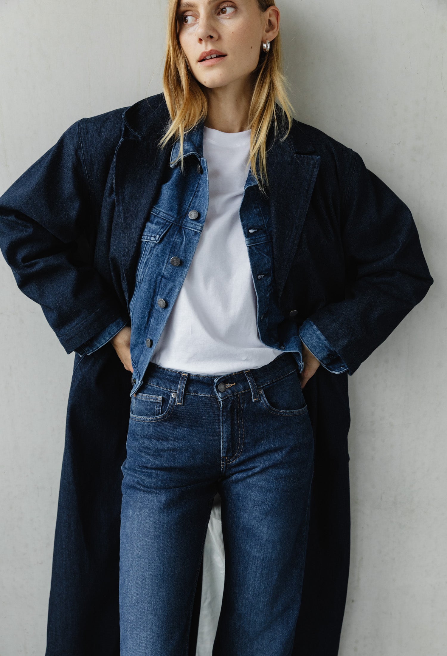 Model wearing layered blue denim jacket and jeans ensemble with a white T-shirt, styled with a long dark coat.
