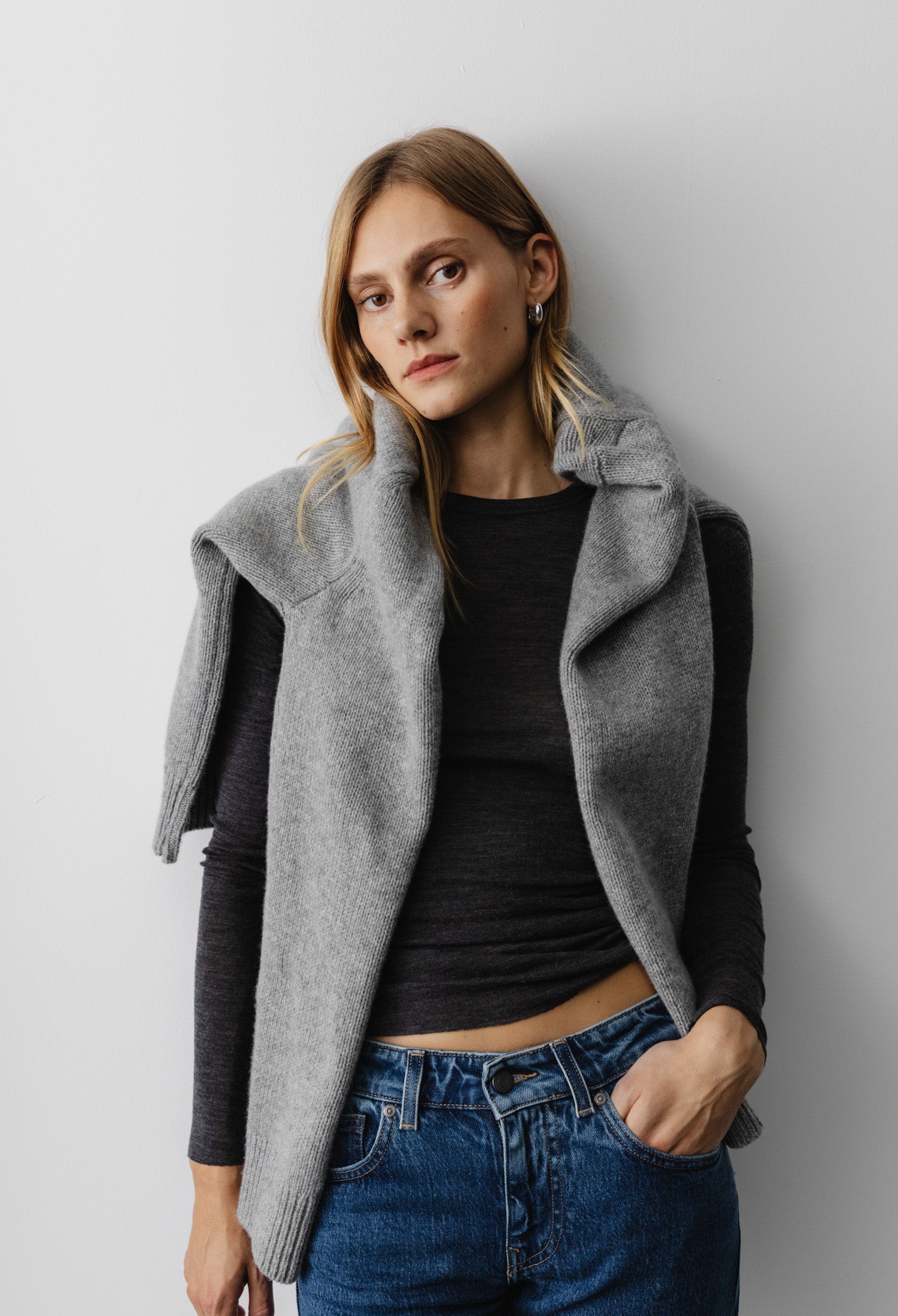 Casual fashion look featuring a gray knit sweater draped over shoulders, black long-sleeve top, and blue denim jeans.