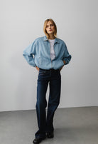 Woman in light blue, oversized denim jacket with dark blue, wide-leg jeans and black shoes, standing against gray wall.
