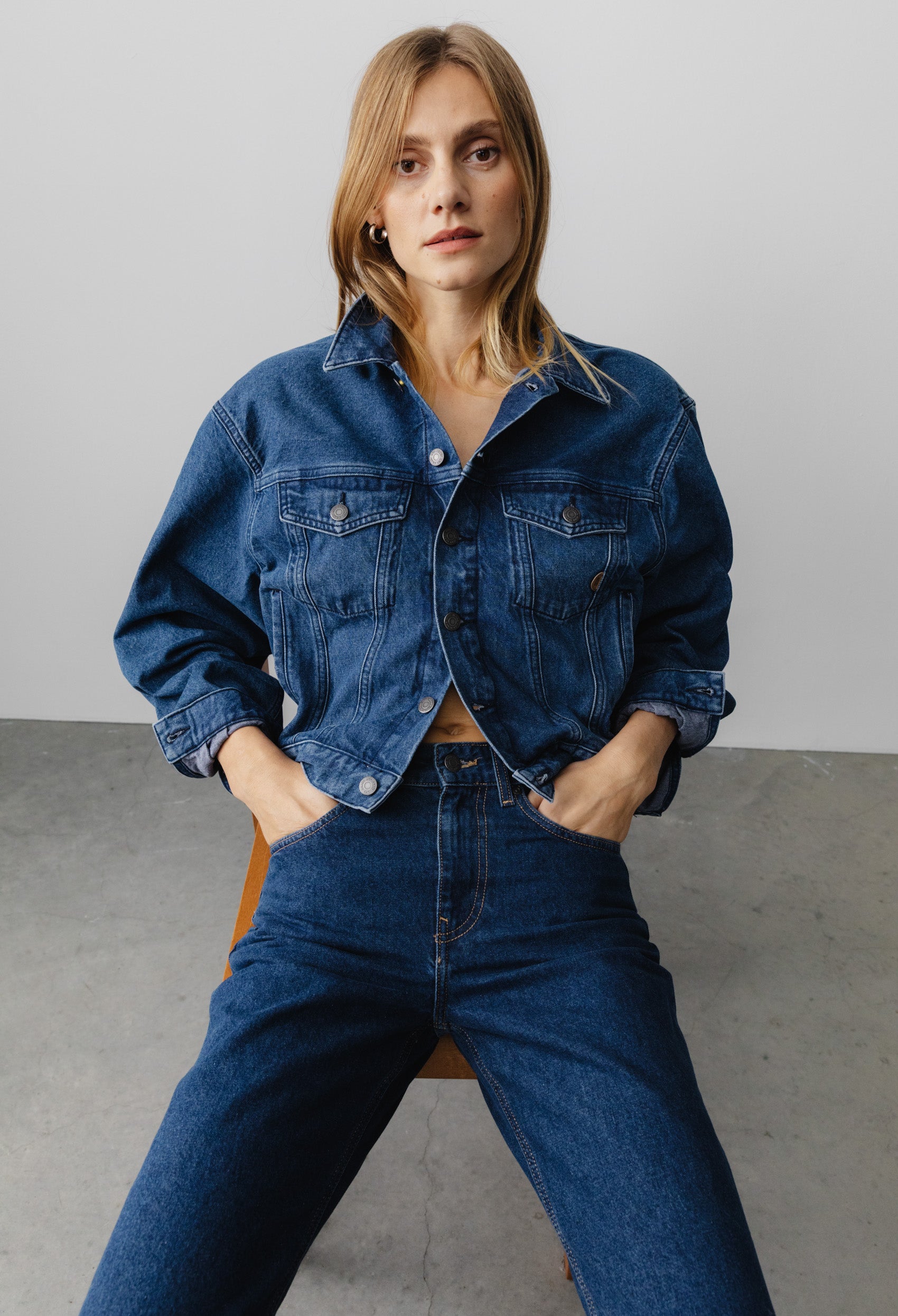 Woman wearing a classic blue denim jacket with matching high-waisted jeans, sitting on a wooden chair against a plain backdrop.
