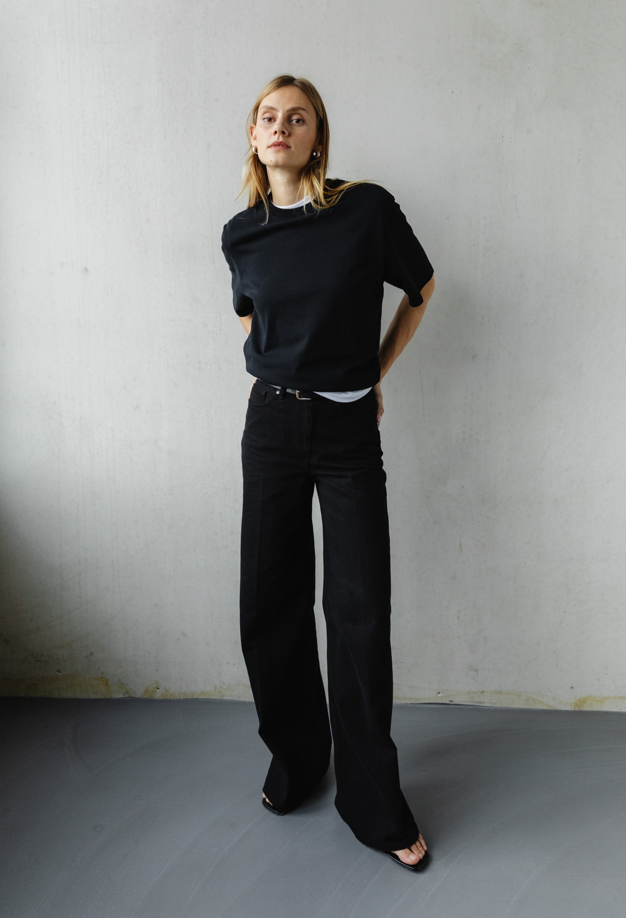 A woman models a minimalist outfit of a black t-shirt and wide-leg black trousers in a neutral indoor setting.