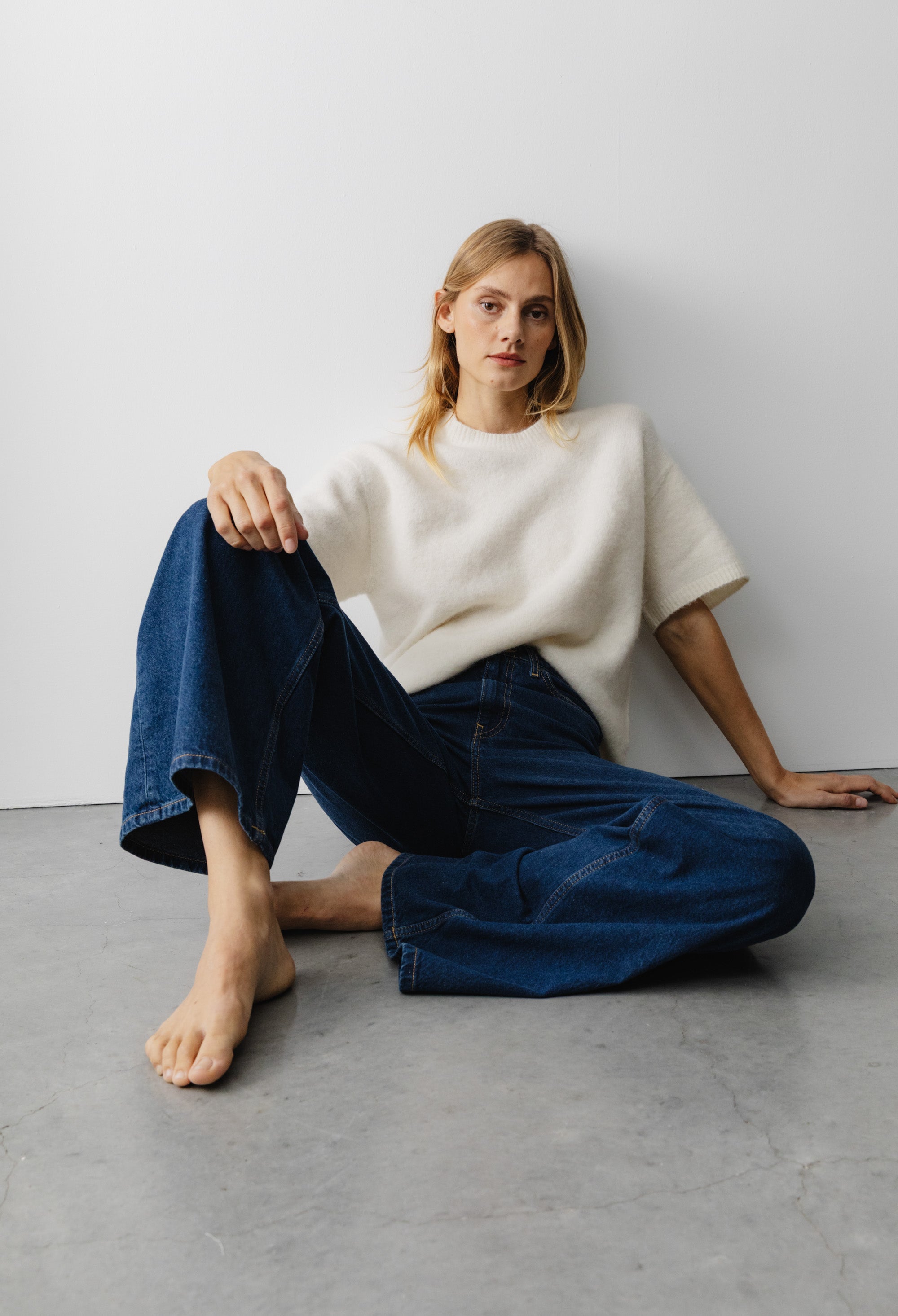 Casual fashion look featuring wide-leg dark blue jeans and soft white short-sleeve sweater on minimalist concrete floor.