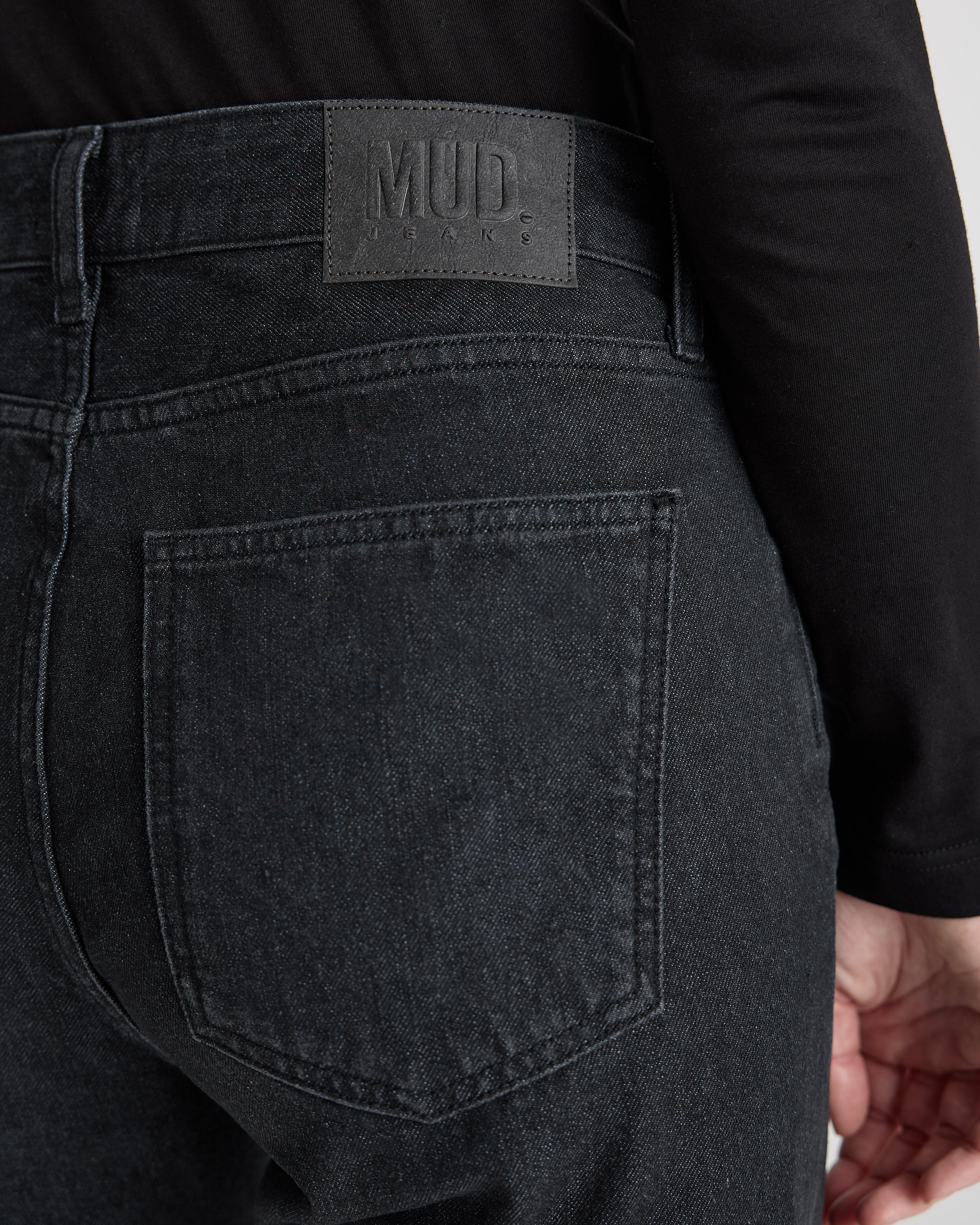 Close-up of MUD Jeans featuring sustainable dark gray denim, showcasing back pocket and leather label, worn with black top.