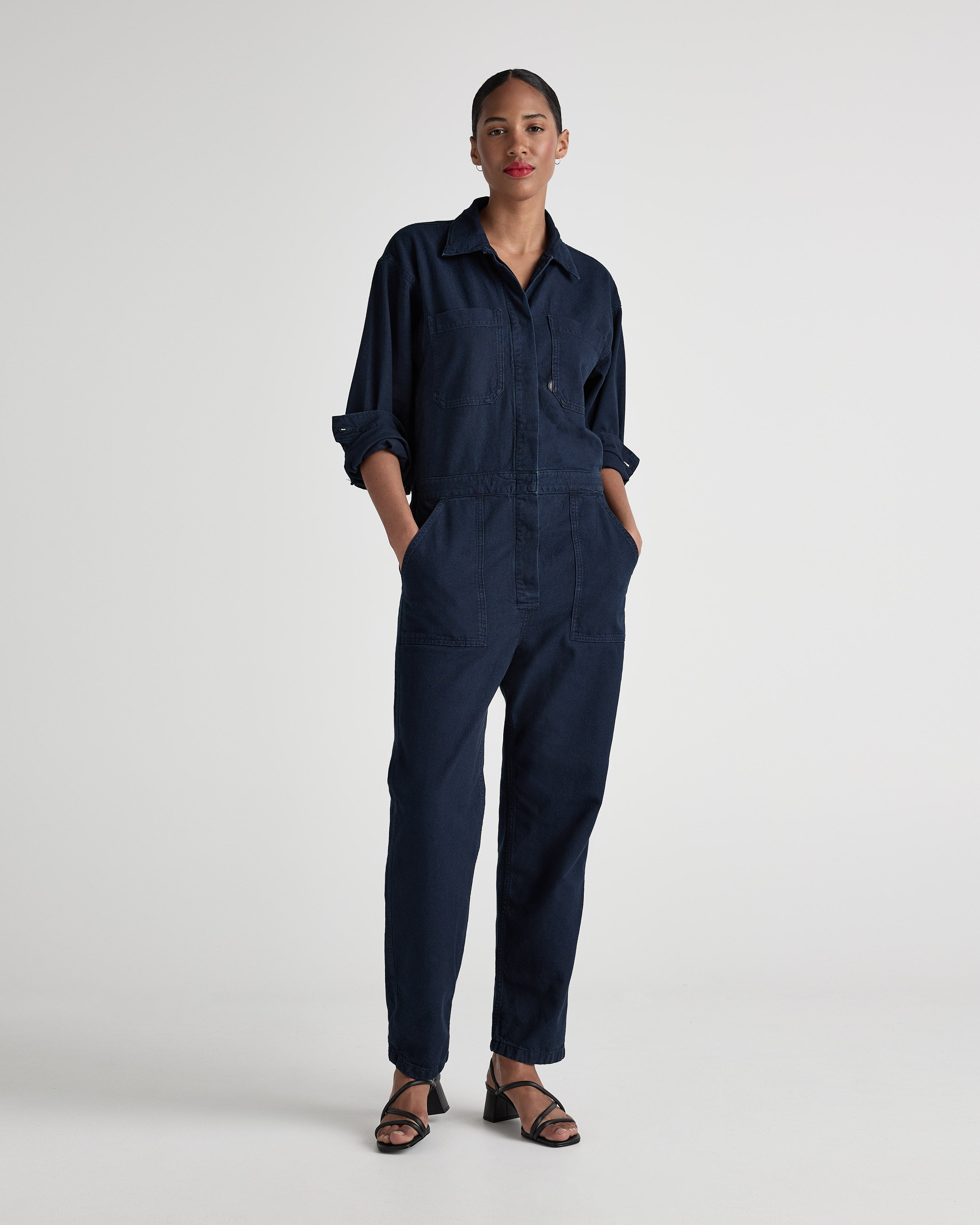Navy blue denim jumpsuit with long sleeves and button-front design, modeled with confidence and paired with black sandals.