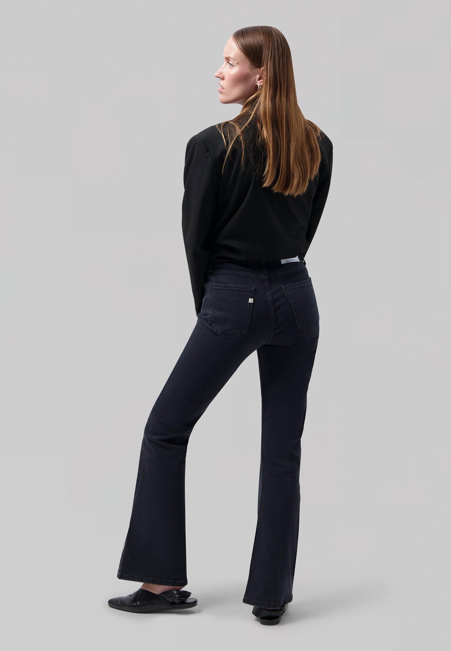 Woman in black long-sleeve top and dark blue flared jeans facing sideways, highlighting sleek, modern fashion style.
