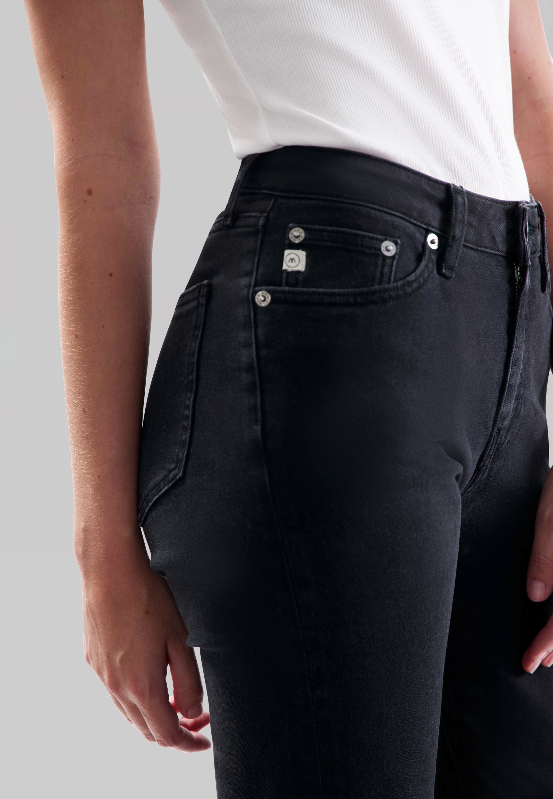 Close-up of black denim jeans with silver buttons, high-waisted fit, and white ribbed tank top tucked in, side view.